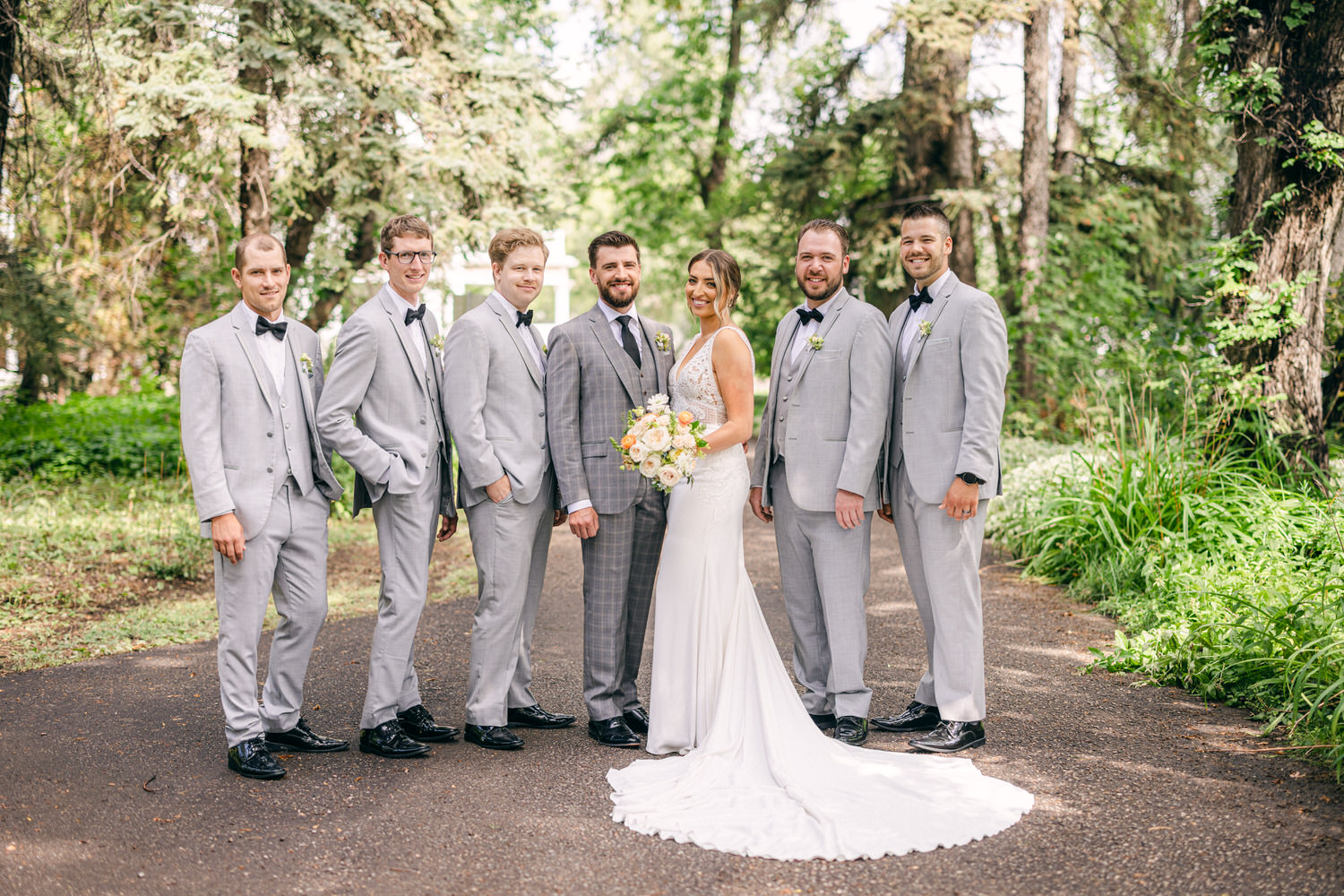 A bride in a beautiful white wedding dress stands in the center of a group of seven groomsmen, all dressed in gray suits, amidst a lush green landscape.