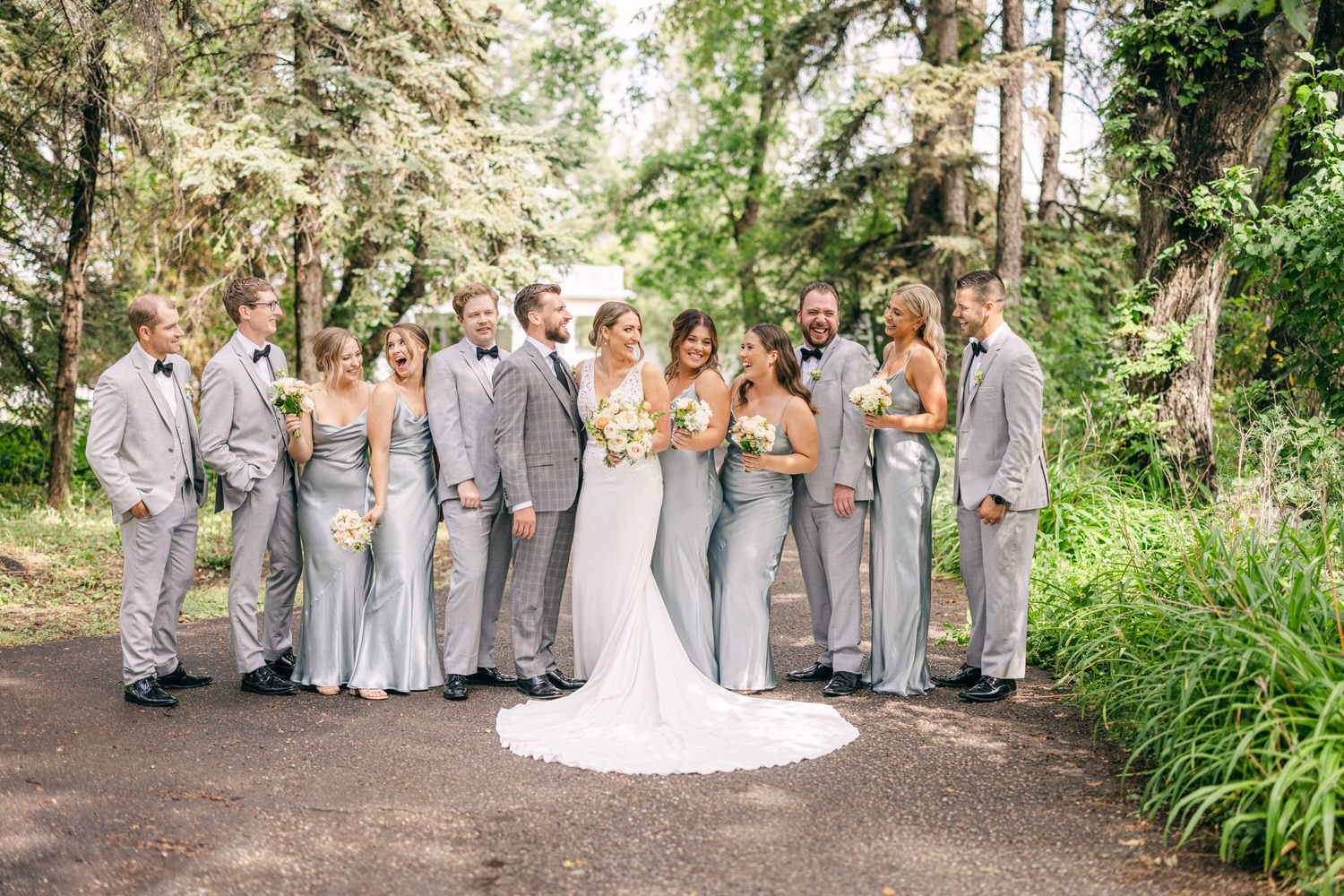 A joyful wedding party posing in a picturesque outdoor setting, featuring a bride and groom surrounded by their happy bridal party in silver attire, vibrant greenery in the background.