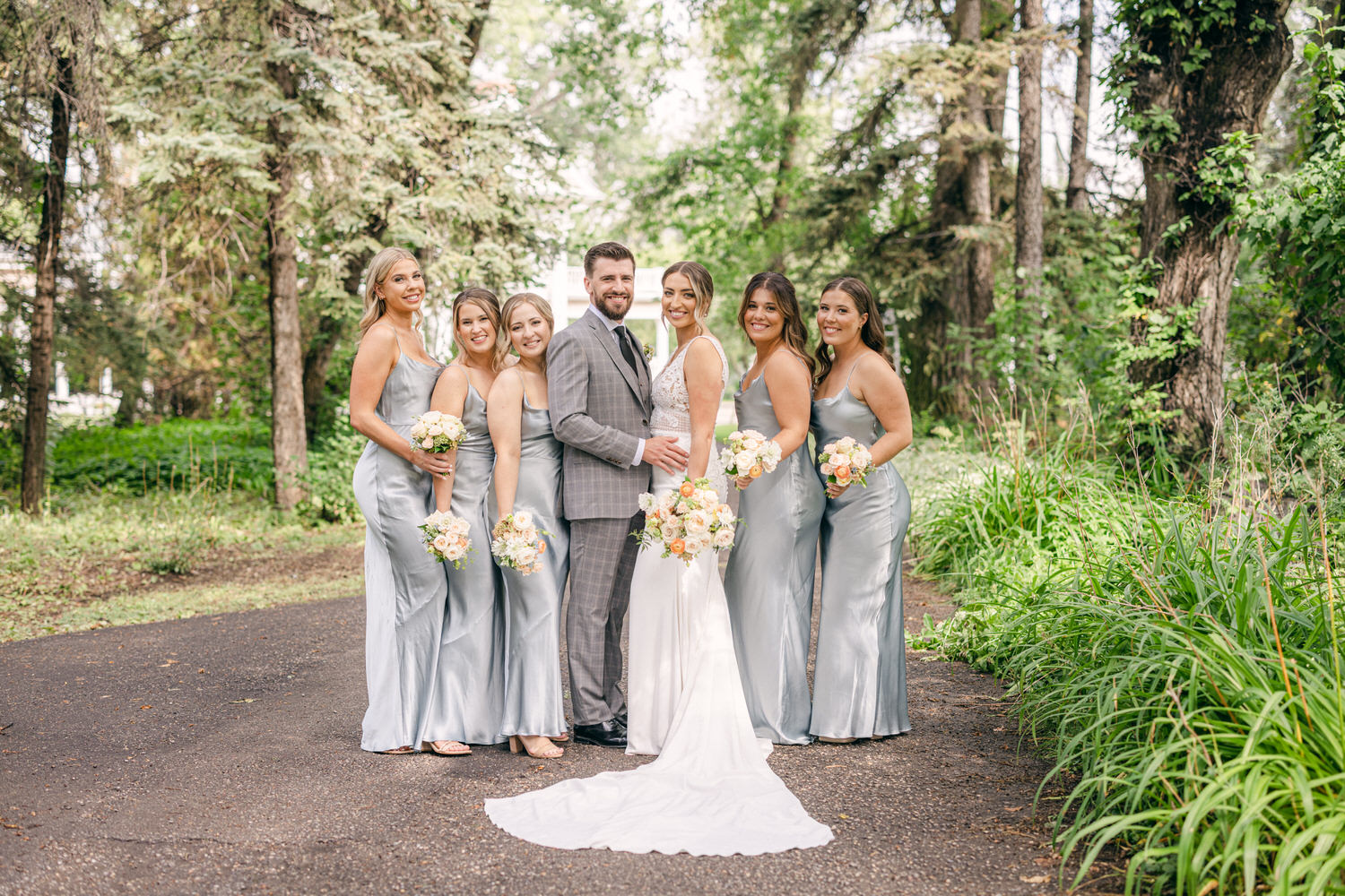 A joyful bride and her bridesmaids, all wearing elegant silver dresses, pose together in a lush green forest with floral bouquets.