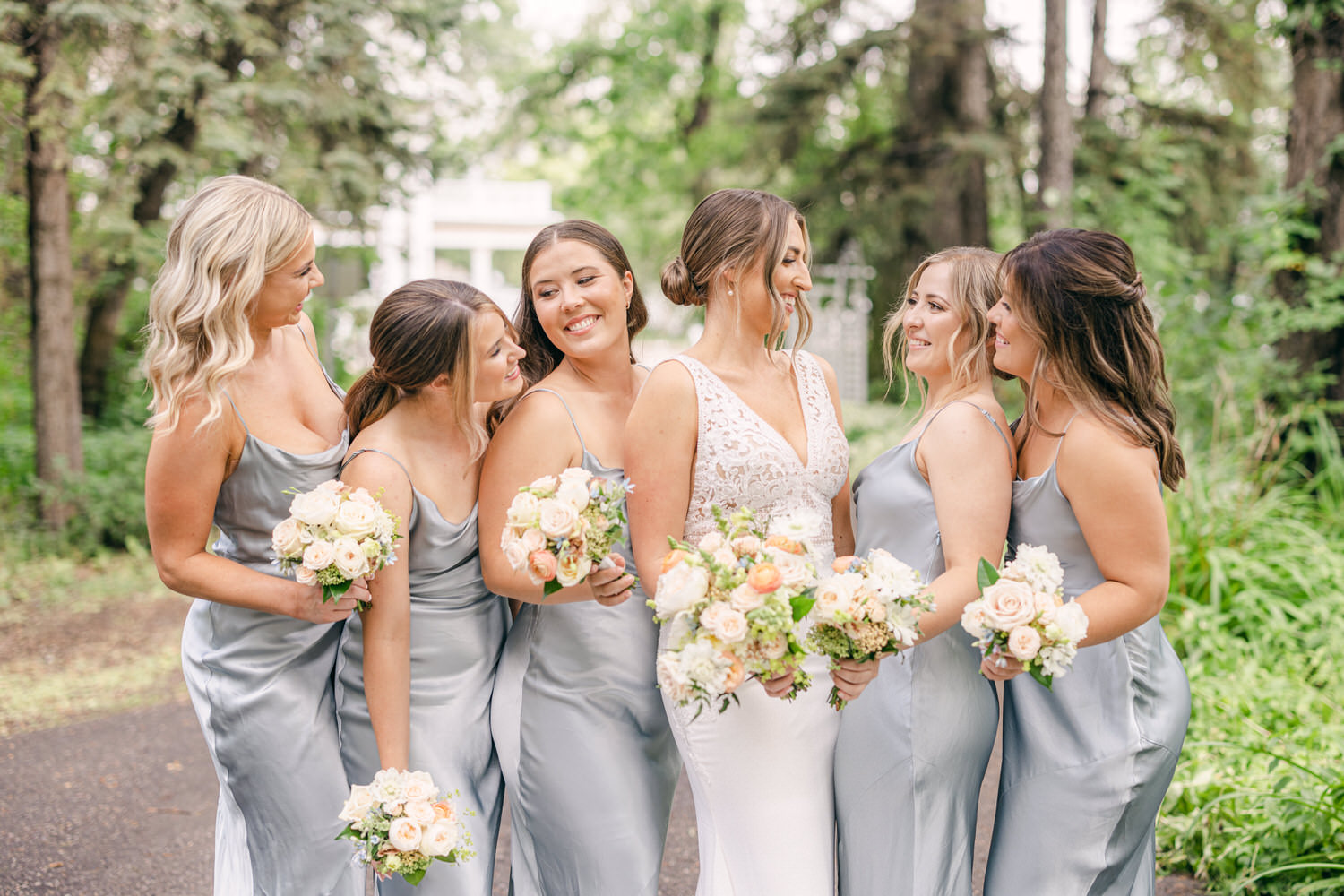 A joyful group of bridesmaids in silver satin dresses, admiring the bride who wears a stunning lace and satin gown, surrounded by lush greenery.