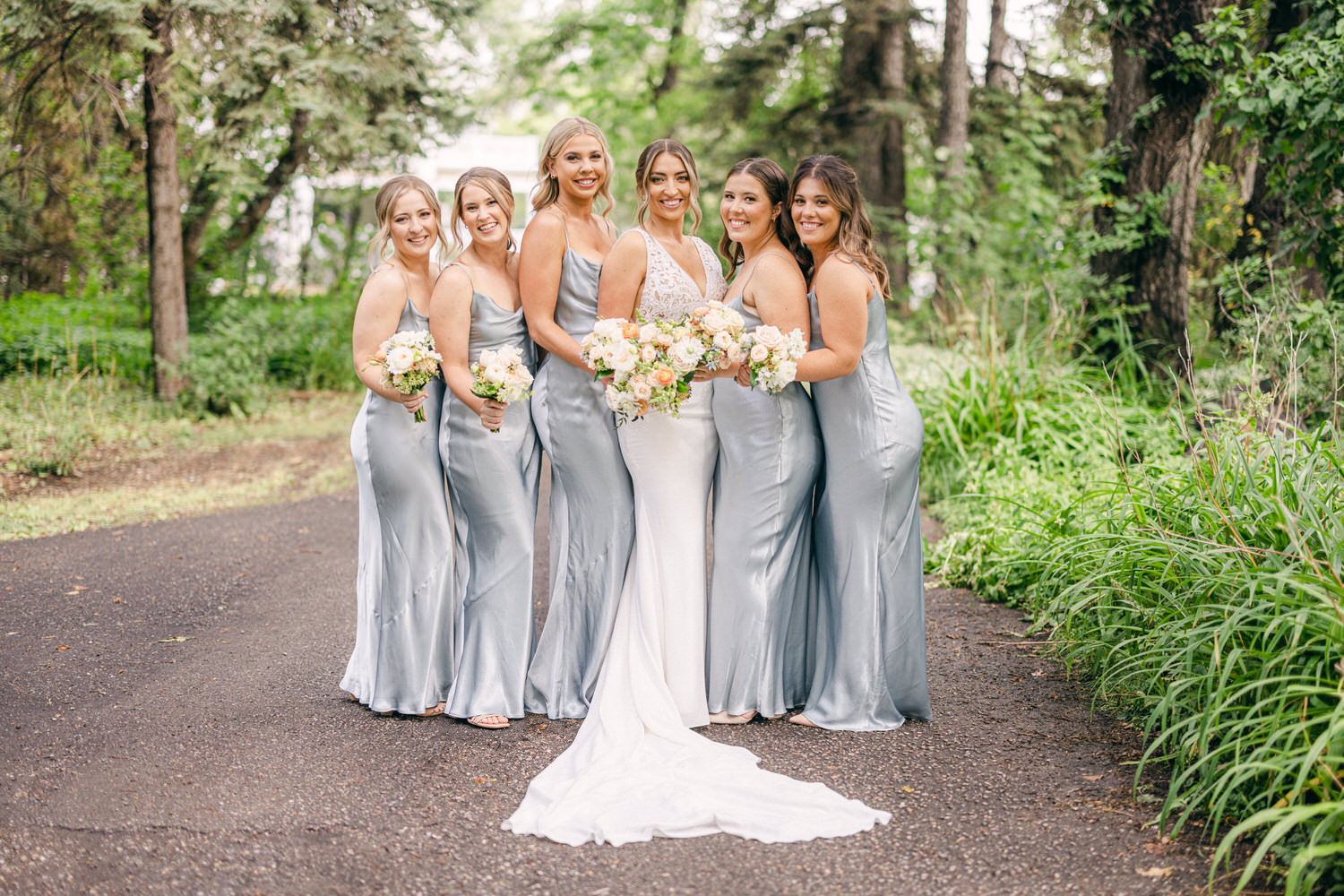 A joyful bridal party poses together on a picturesque path, wearing matching silver gowns and holding bouquets of soft flowers, with a lush green backdrop.