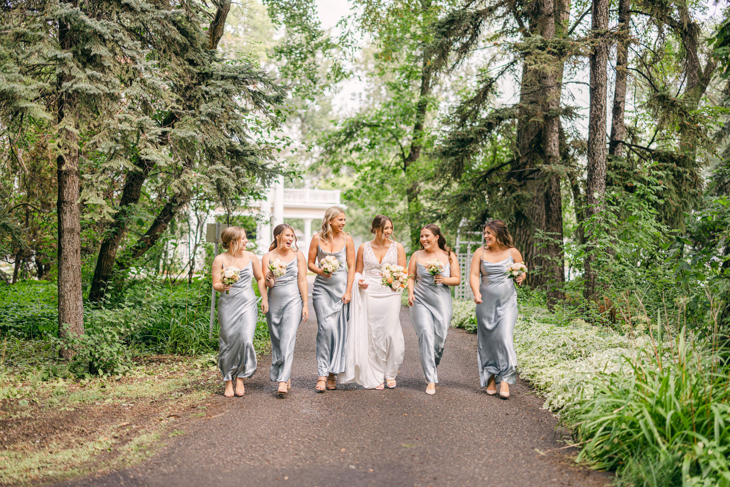 A joyful bridal party walks down a scenic path, wearing elegant silver dresses and holding floral bouquets, amidst lush greenery.