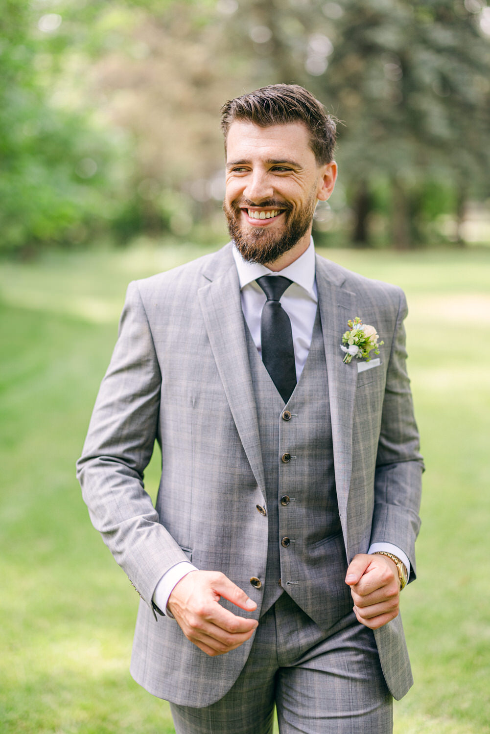 Stylish Man in Gray Suit::A confident man in a gray plaid suit and black tie smiles while walking through a lush green park, showcasing a boutonniere on his lapel.