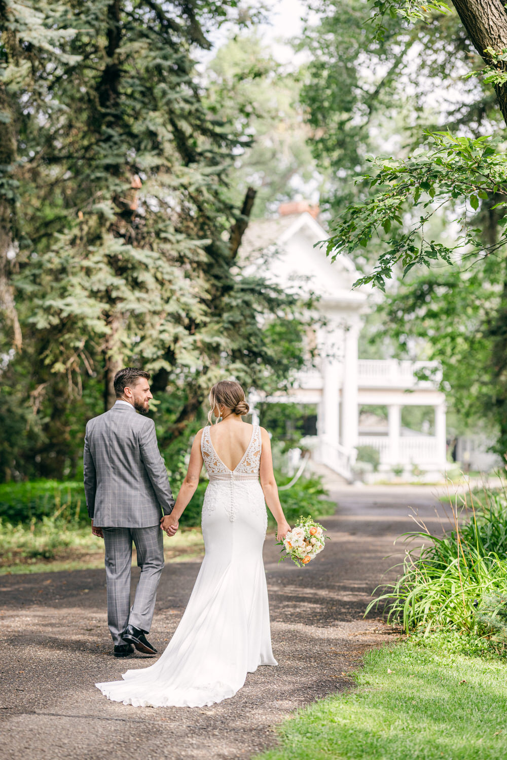 A couple walks hand in hand down a scenic path, surrounded by lush greenery and a charming white house in the background, celebrating their wedding day.