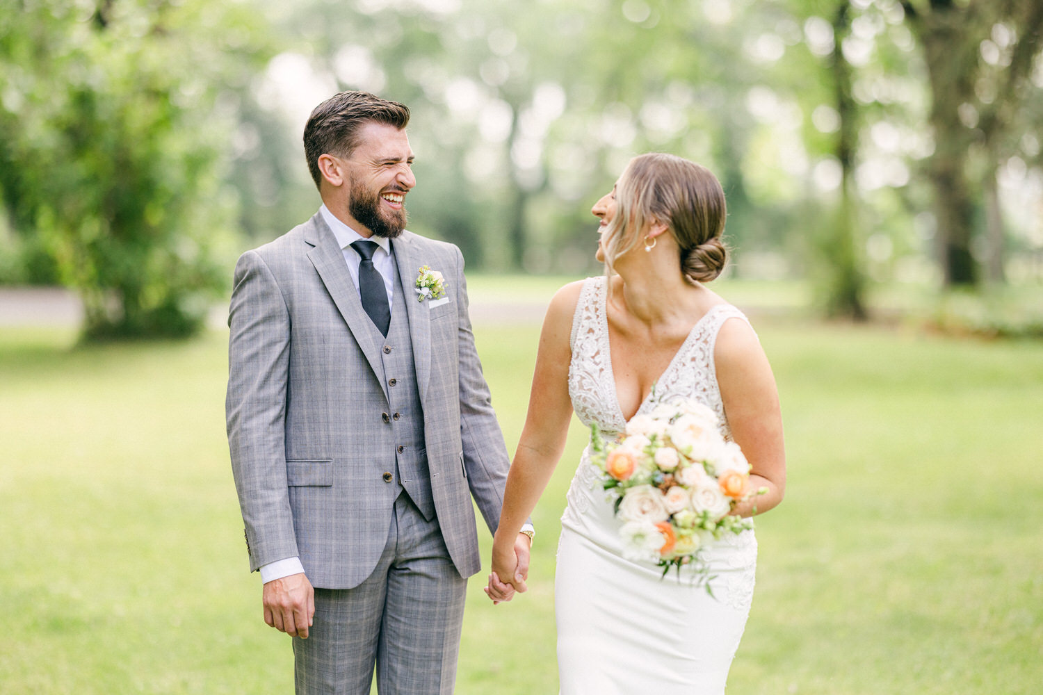 A couple laughing together while holding hands outdoors, surrounded by greenery, capturing a joyful moment on their wedding day.