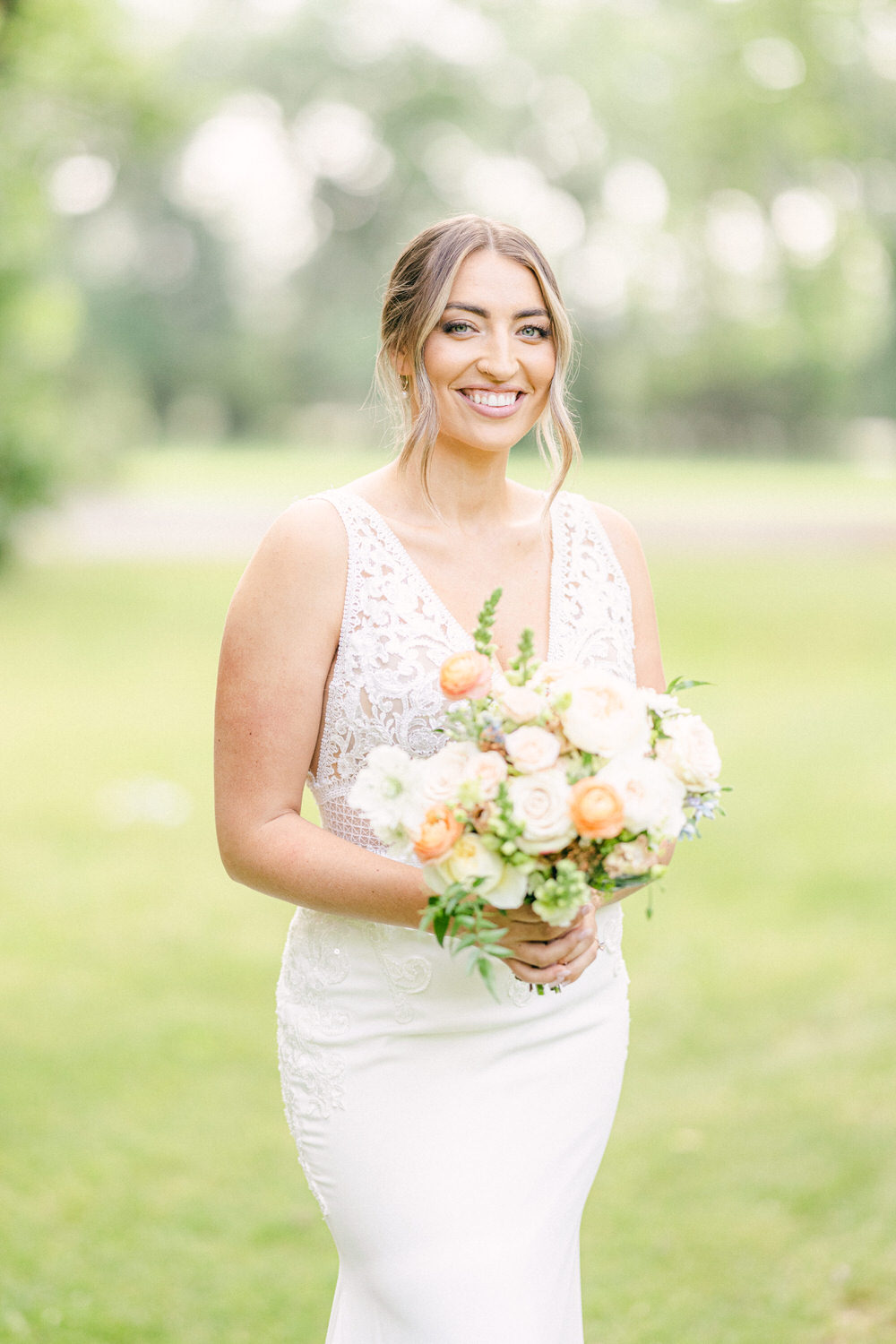 A smiling bride in a sleek white gown holds a beautiful bouquet of pastel flowers, standing in a vibrant green garden.