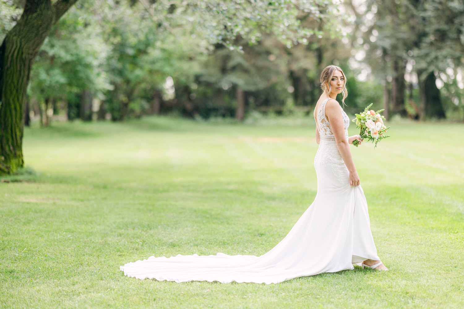 A bride in a fitted white gown walks gracefully in a lush green outdoor setting, holding a bouquet of pastel flowers.