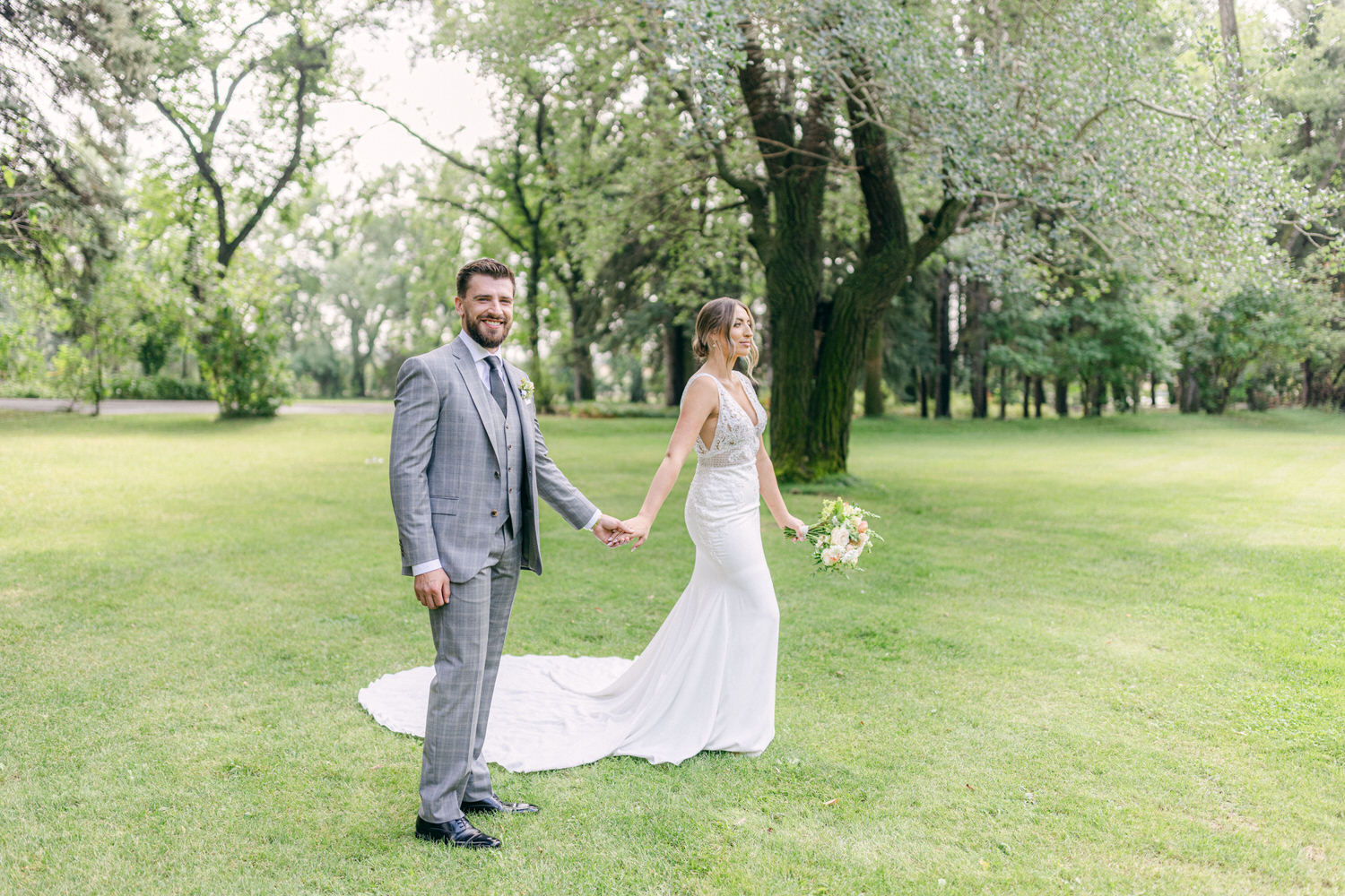 A joyful couple in wedding attire stands together in a lush green park, with the bride holding a bouquet and the groom smiling at the camera.