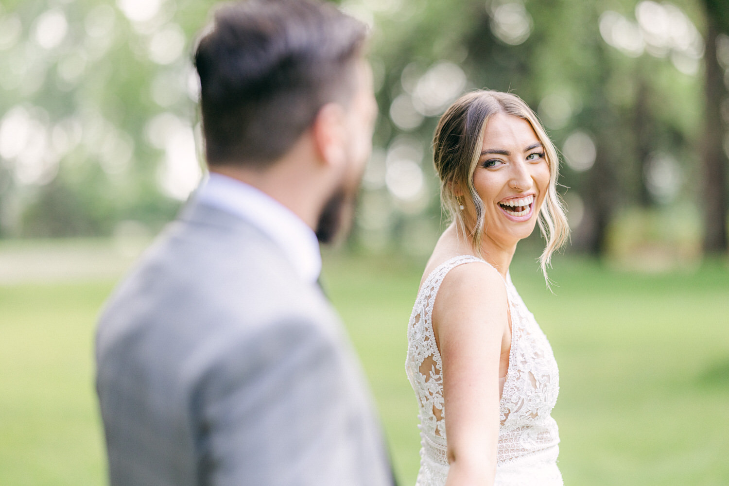 A smiling bride wearing a lace dress turns to look at her partner, surrounded by a blurred green background, capturing a moment of happiness.