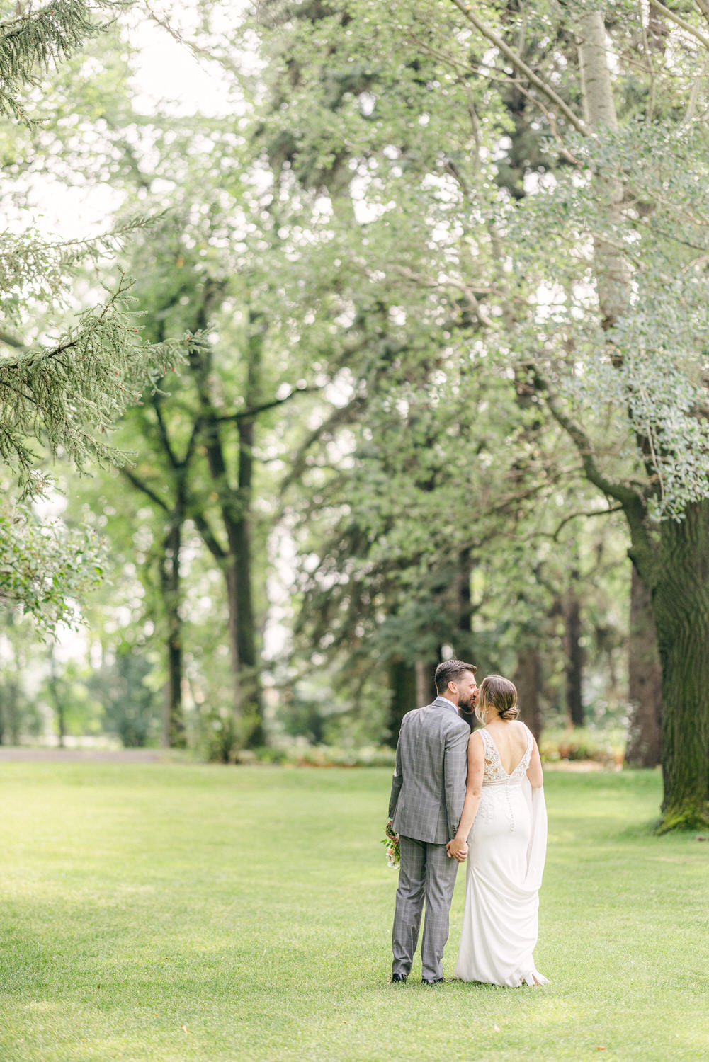 Wedding Couple in a Serene Park:::A newlywed couple holding hands and walking away, surrounded by lush greenery in a tranquil park setting.