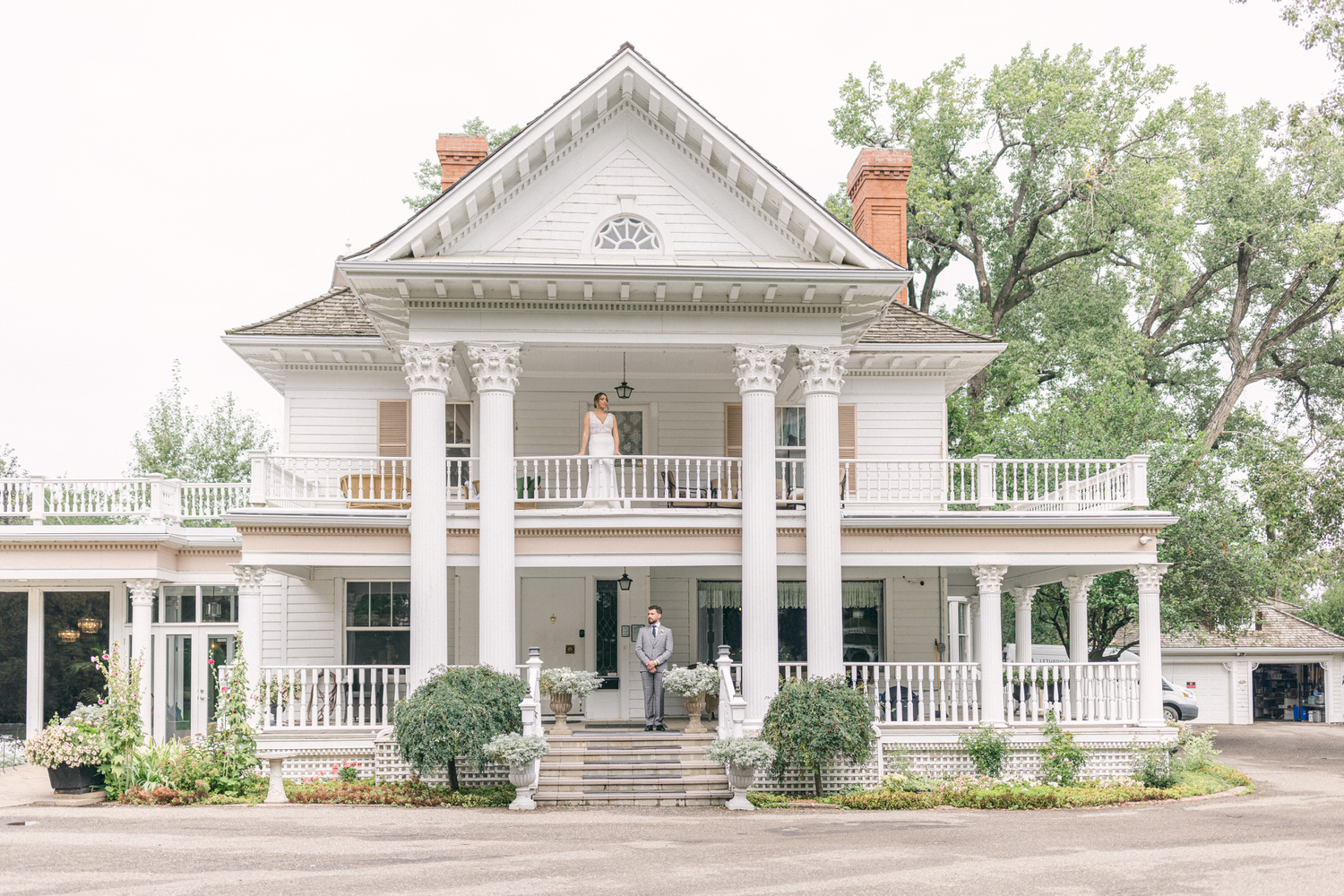 A bride in a white dress stands on a balcony of a classic mansion while a groom waits below on the steps, surrounded by lush greenery and potted plants.