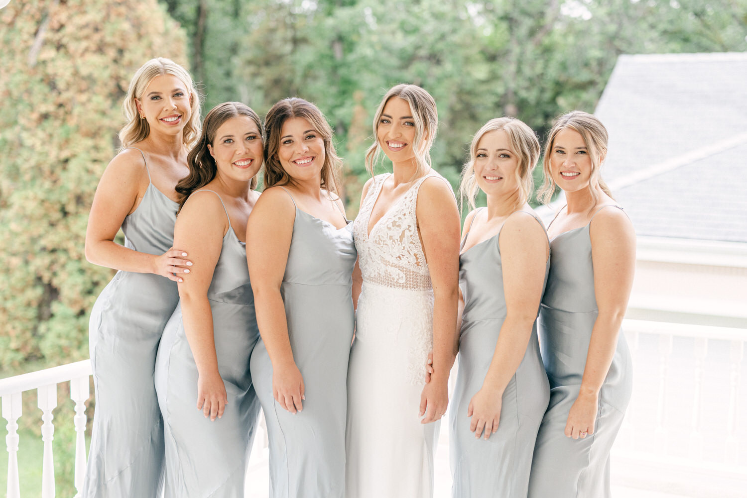 A joyful bride poses with her five bridesmaids, all wearing elegant gray dresses, against a lush green backdrop.