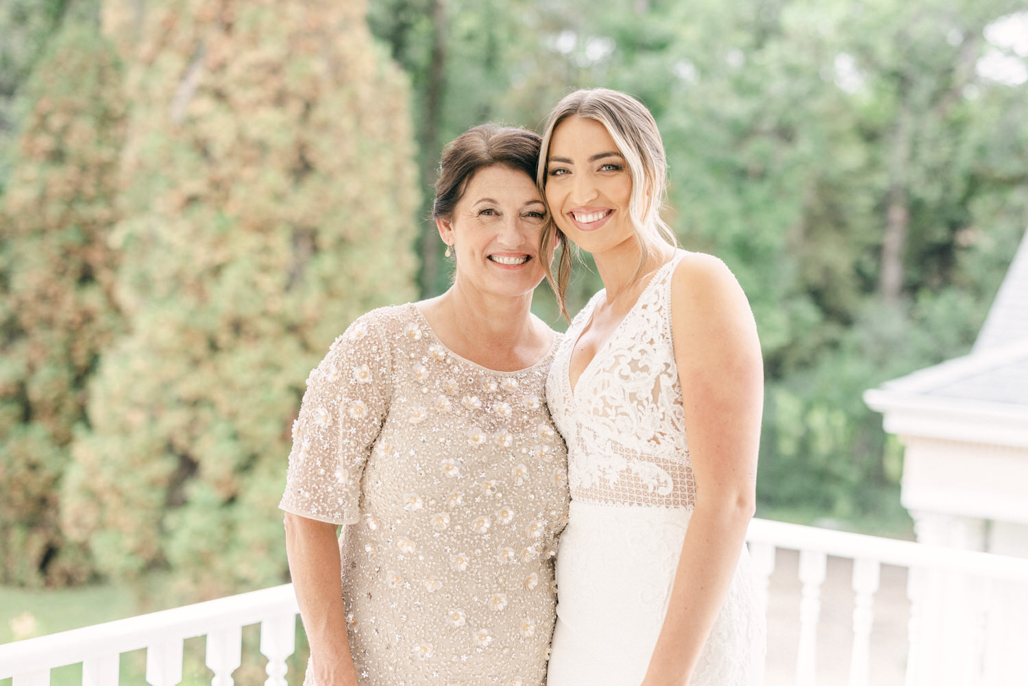 A smiling mother and daughter pose together outdoors, with greenery in the background, both wearing elegant outfits.