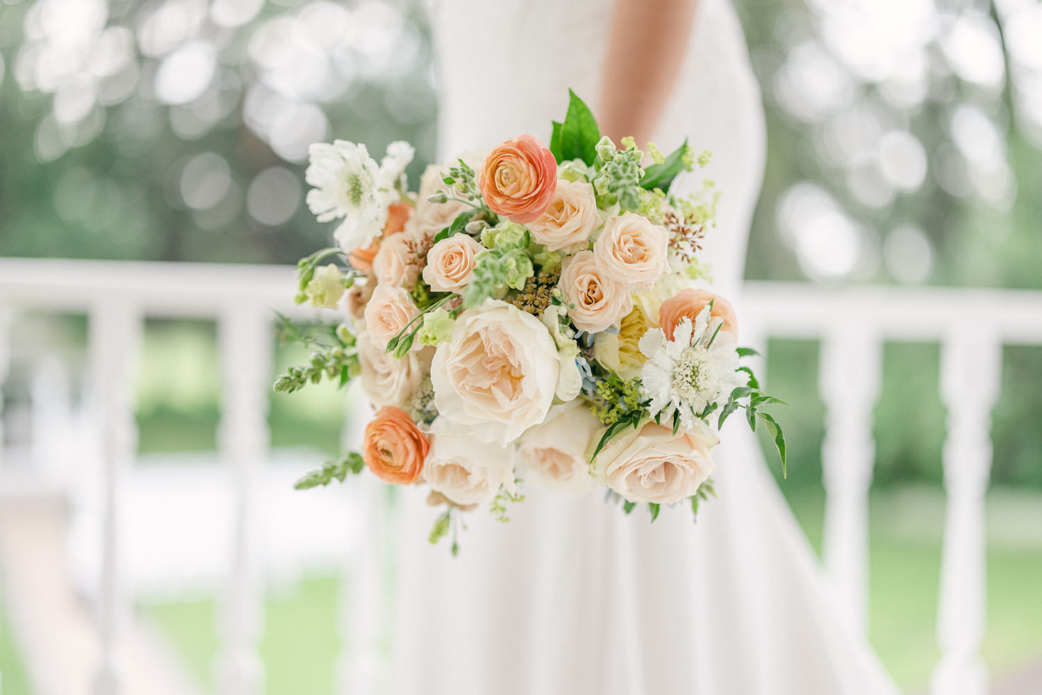 A close-up of a bridal bouquet featuring soft pastel roses, ranunculus, and greenery, held by a bride against a blurred outdoor backdrop.