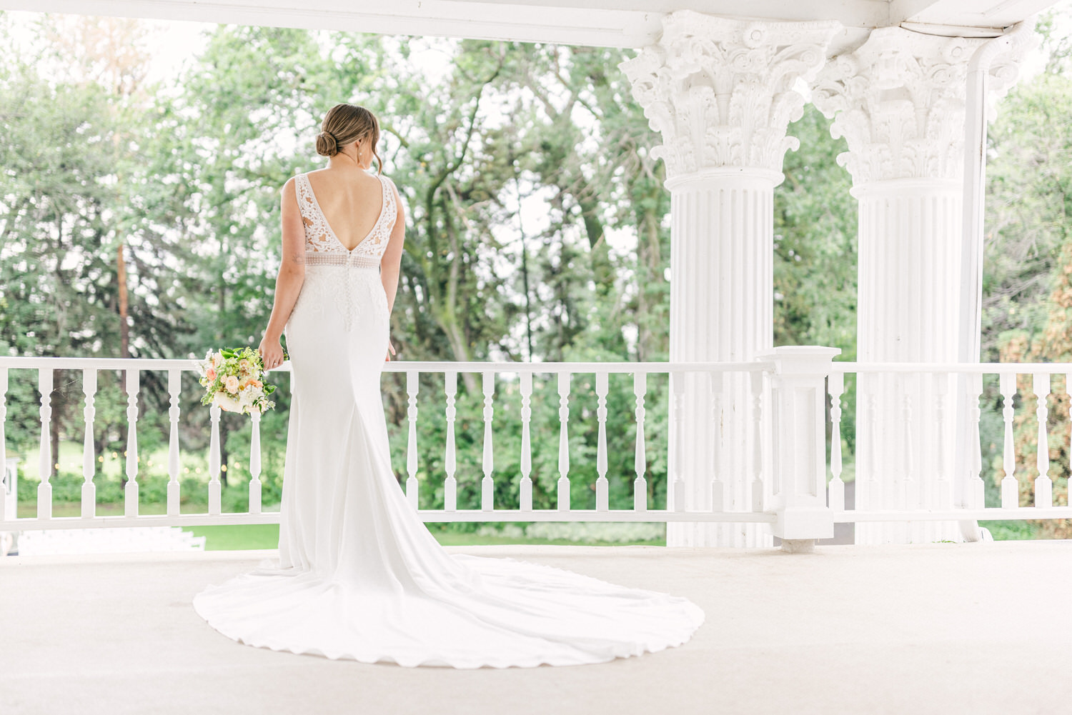 A bride stands gracefully on a porch, showcasing her elegant white gown and bouquet against a lush green backdrop.