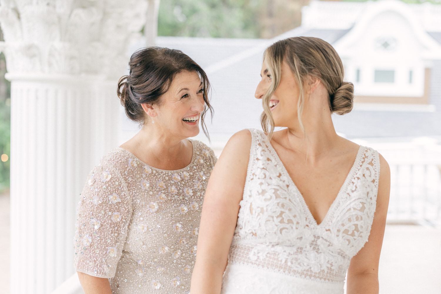 A smiling mother and bride share a joyful moment together in elegant attire, surrounded by a beautifully detailed indoor setting.