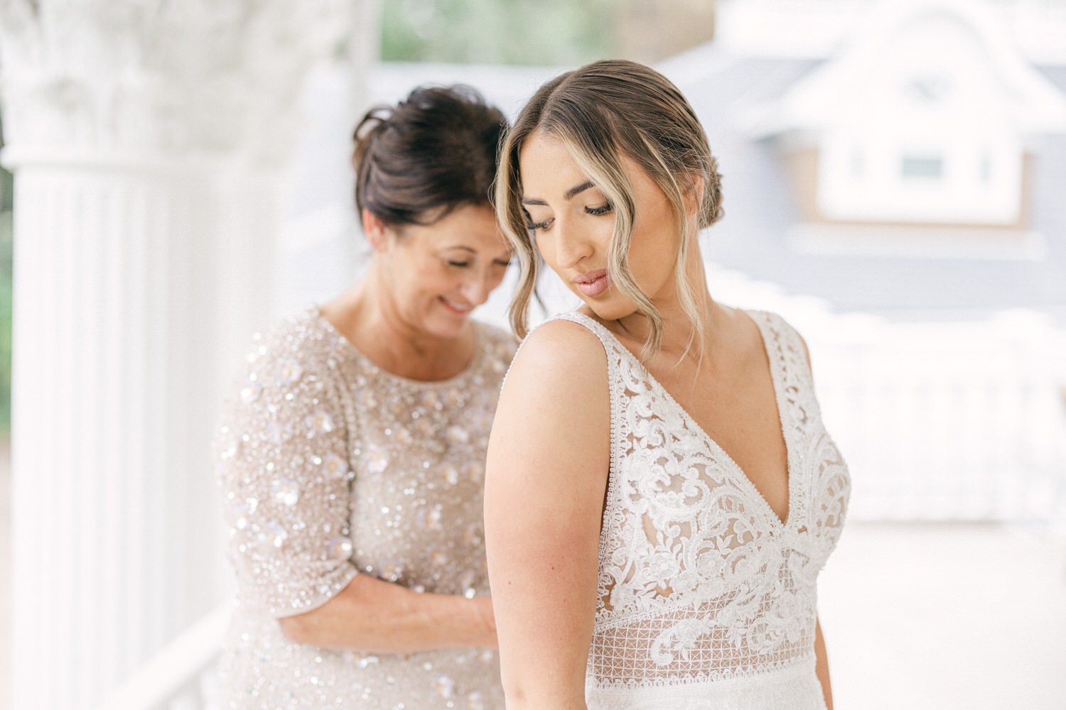 A bride in a delicate white gown poses while her mother adjusts her dress, showcasing a tender moment before the wedding.