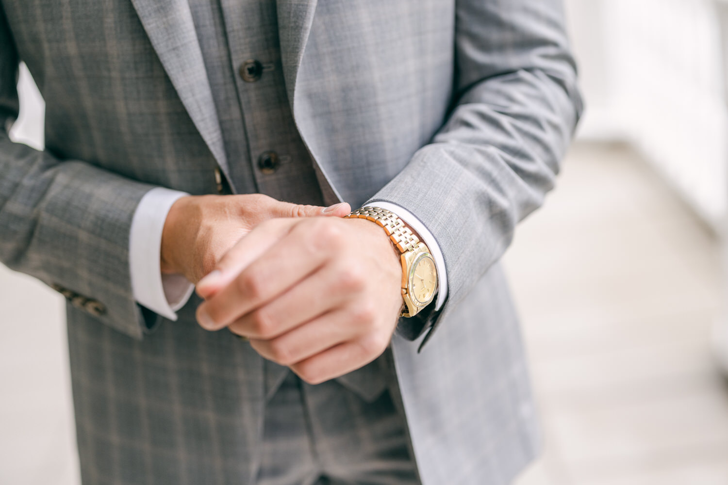 Close-up of a man's hands adjusting his wristwatch while wearing a gray plaid suit.