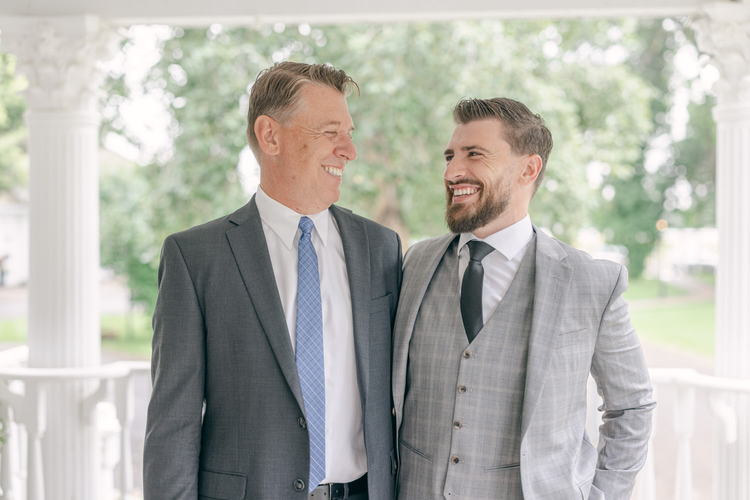 Two men in formal suits share a warm smile and laughter outdoors, surrounded by greenery.