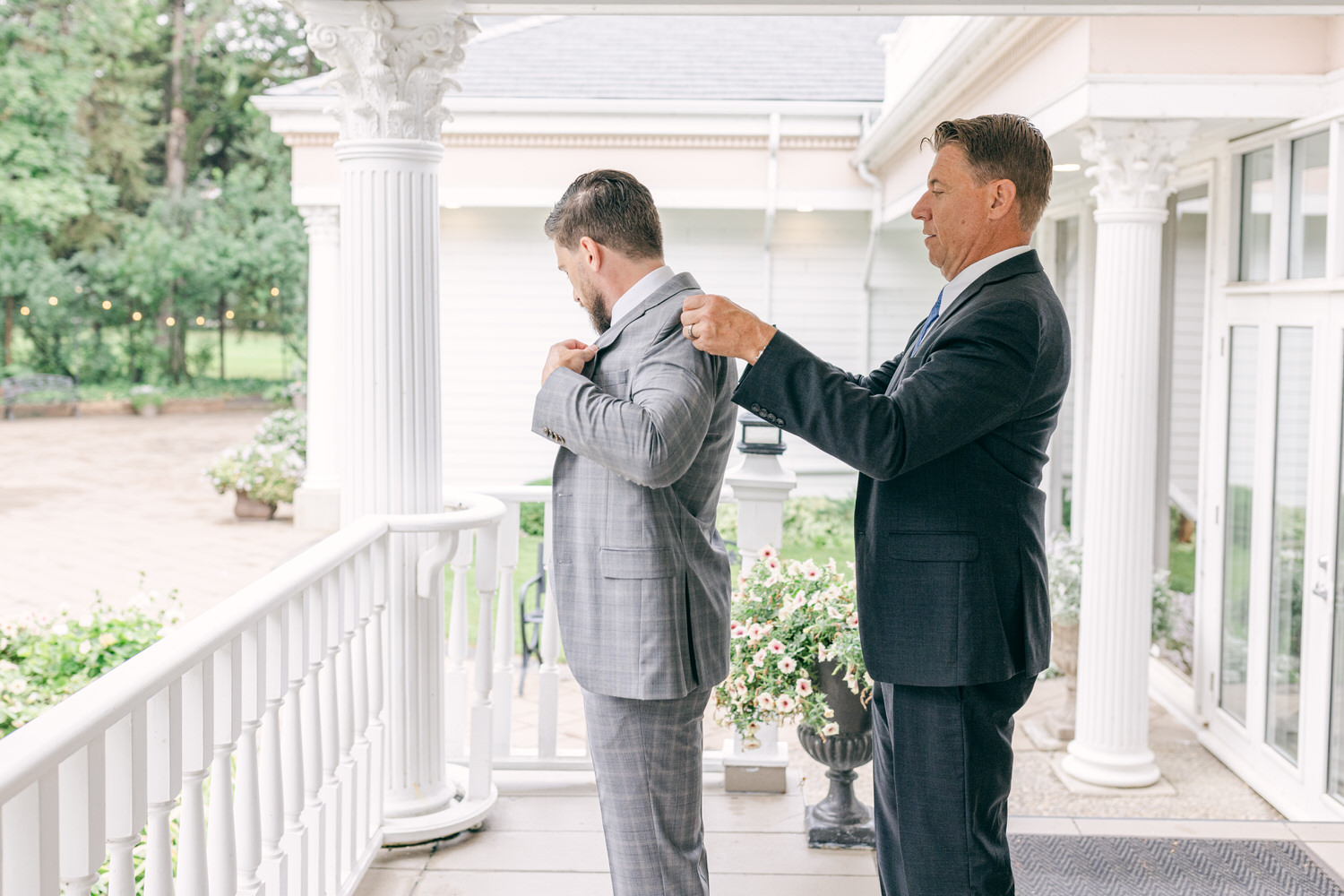 A man in a grey plaid suit receives assistance with his jacket from another man in a black suit, set against a beautiful outdoor venue with greenery and flowers.