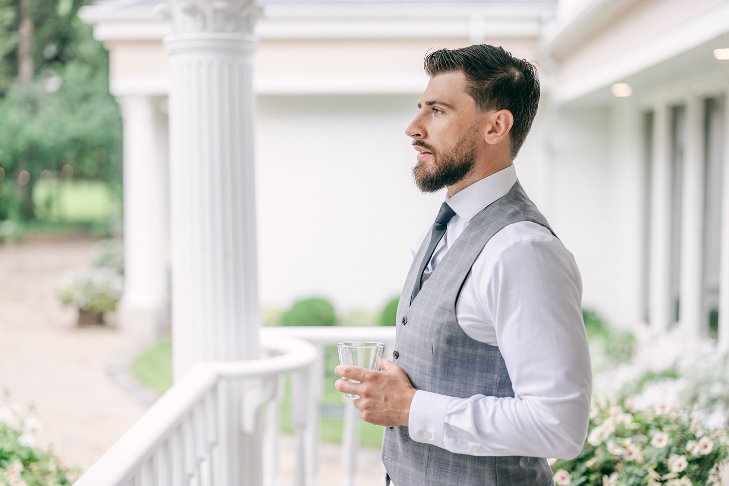 A well-dressed man with a beard stands outdoors, holding a glass, wearing a grey vest over a white shirt and black tie, with a soft-focus garden in the background.