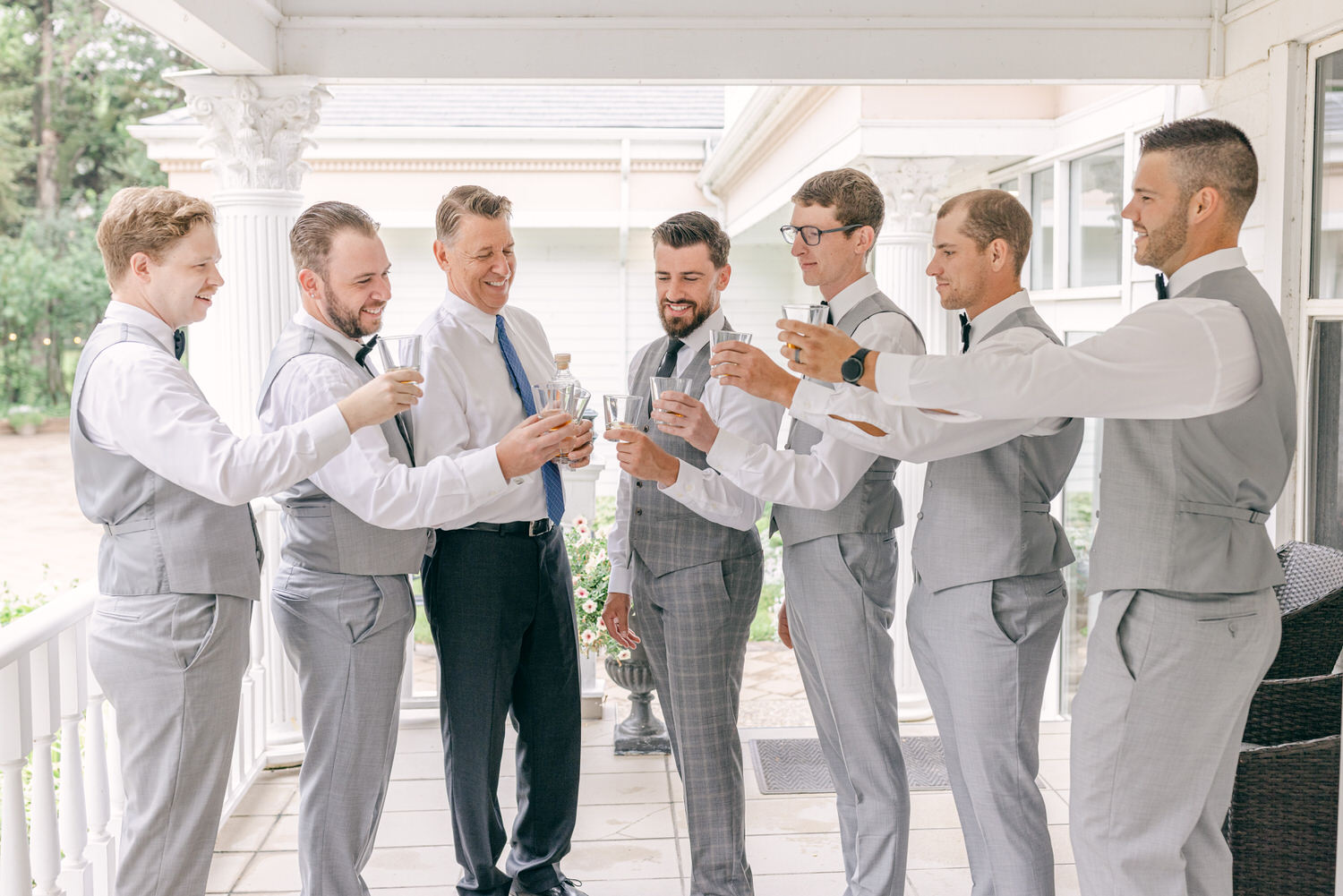 A group of seven men in gray suits toast with clear glasses, celebrating in a light and cheerful setting.