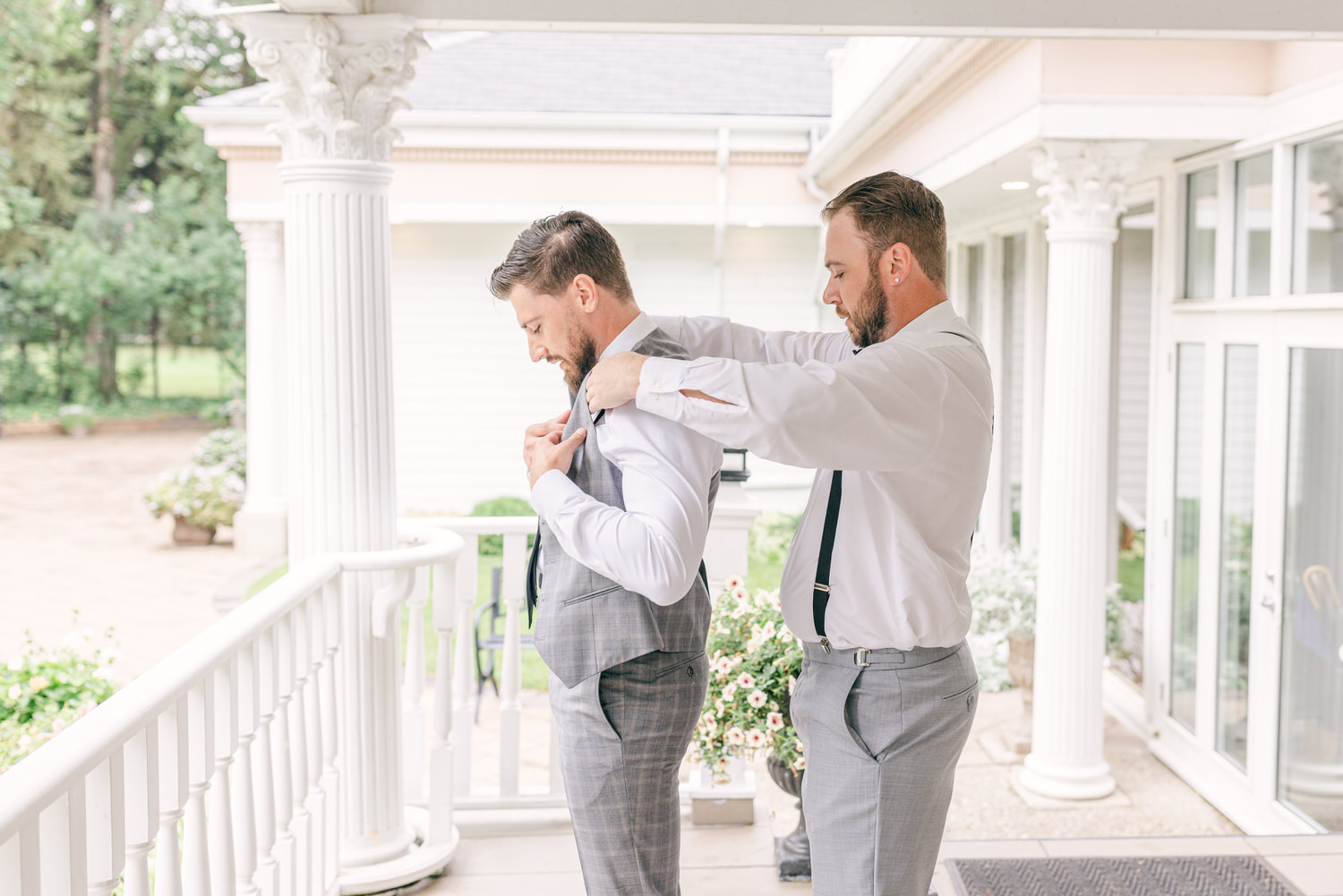 Two men, one in a grey suit and another in a white shirt, help each other with wedding attire adjustments on a porch surrounded by greenery.