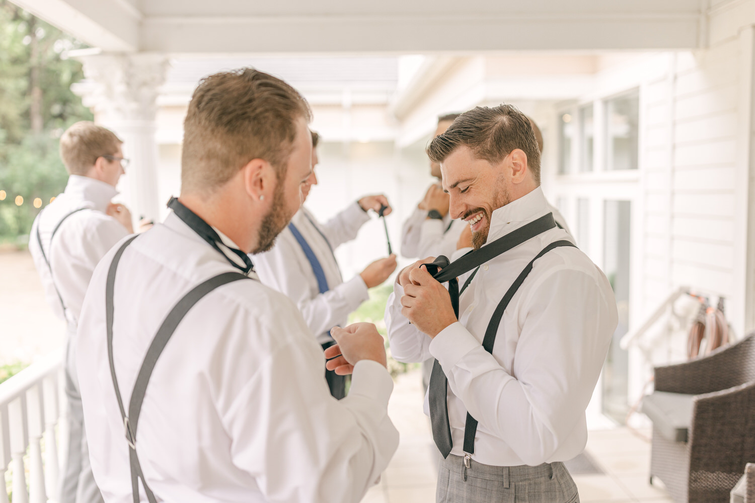 Groom and groomsmen adjusting their ties and suspenders while getting ready for the wedding in a bright, relaxed setting.