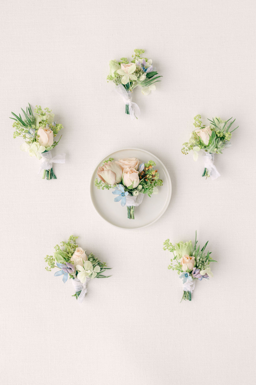 A delicate arrangement of five floral boutonnieres featuring pale roses and assorted greenery, with one centerpiece boutonniere on a plate, all set against a soft white background.