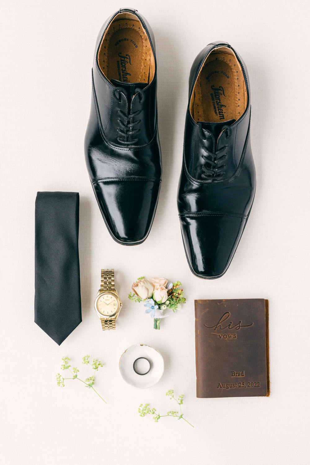 An arrangement of polished black dress shoes, a black tie, a gold watch, a floral boutonniere, a gold ring in a dish, and a leather vow book labeled "his vows" with a name and date on a light background.