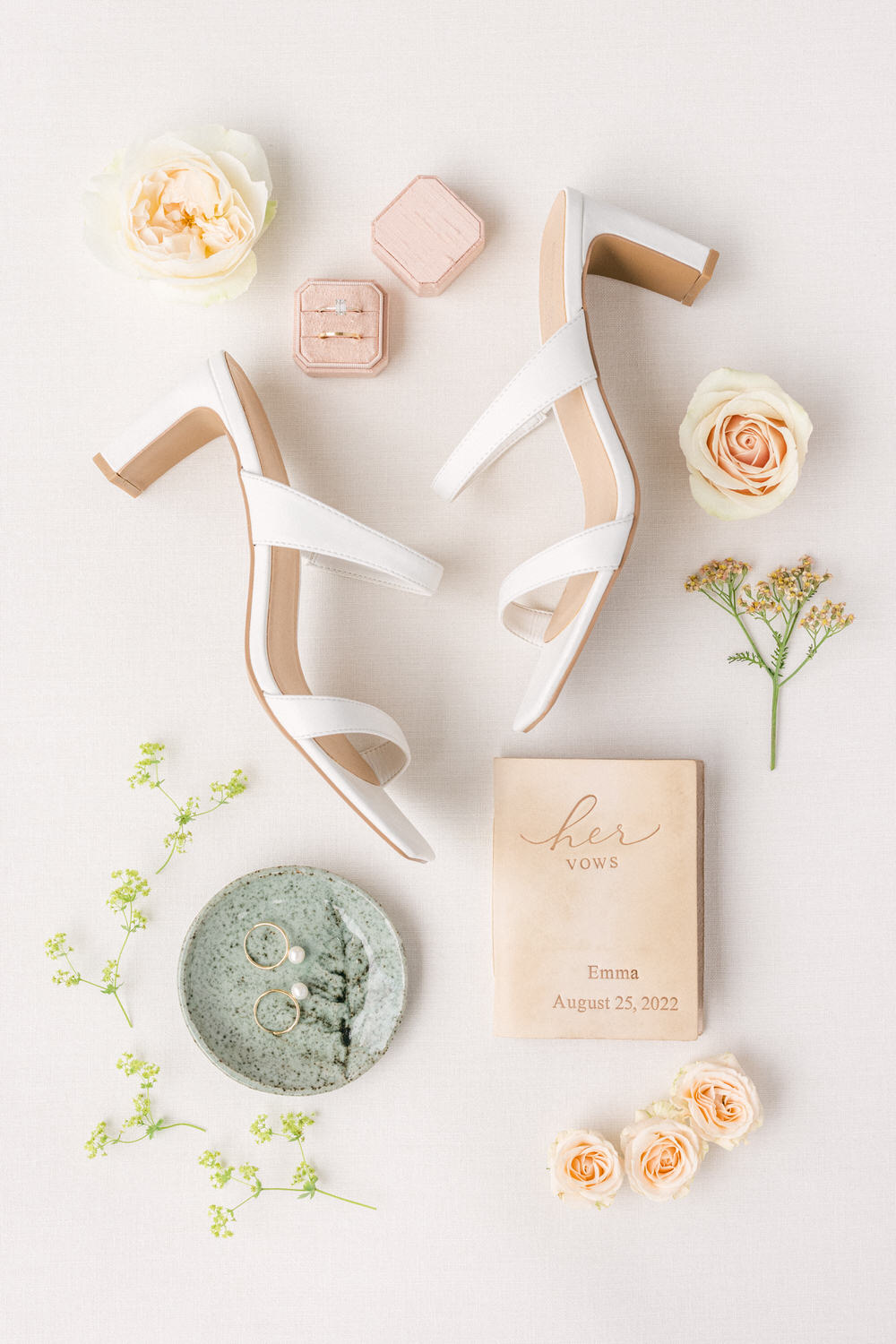 A flat lay of white high-heeled sandals, vow book, floral arrangements, and wedding jewelry on a light fabric background.