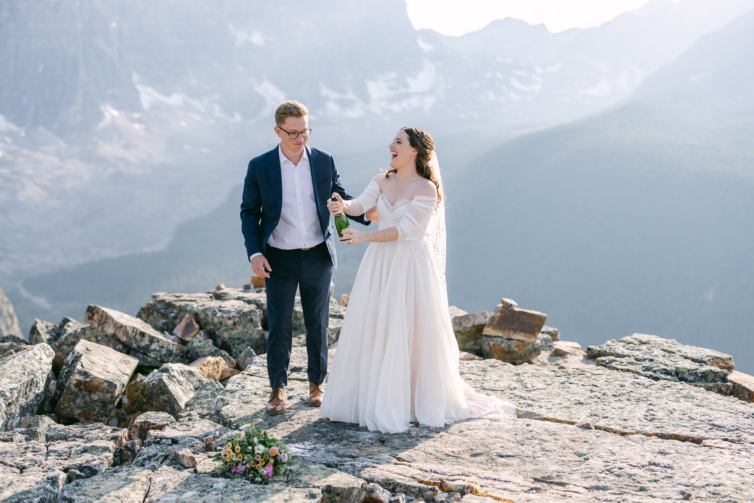 A joyful couple in wedding attire celebrating on a rocky mountain top, surrounded by stunning scenic views. The woman, in a flowing white gown, is popping a champagne bottle, while the man in a suit smiles beside her, with a bouquet of flowers at their feet.