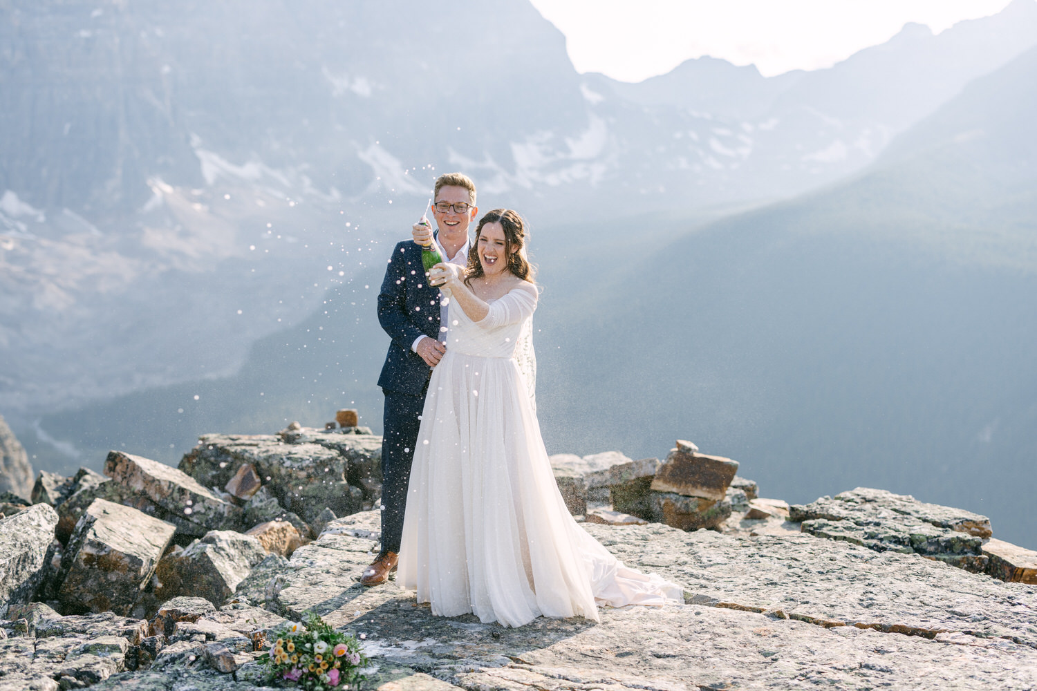 A joyful couple pops champagne on a rocky mountain peak, surrounded by stunning mountain scenery, reflecting their happiness on their special day.