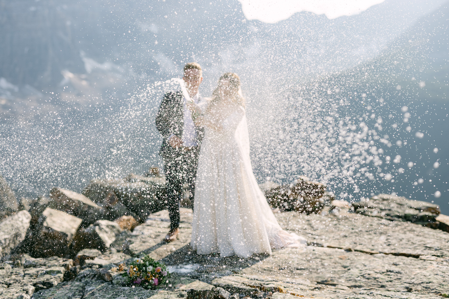 A bride and groom standing on rocky terrain, surrounded by sparkling water droplets in a breathtaking mountainous backdrop.