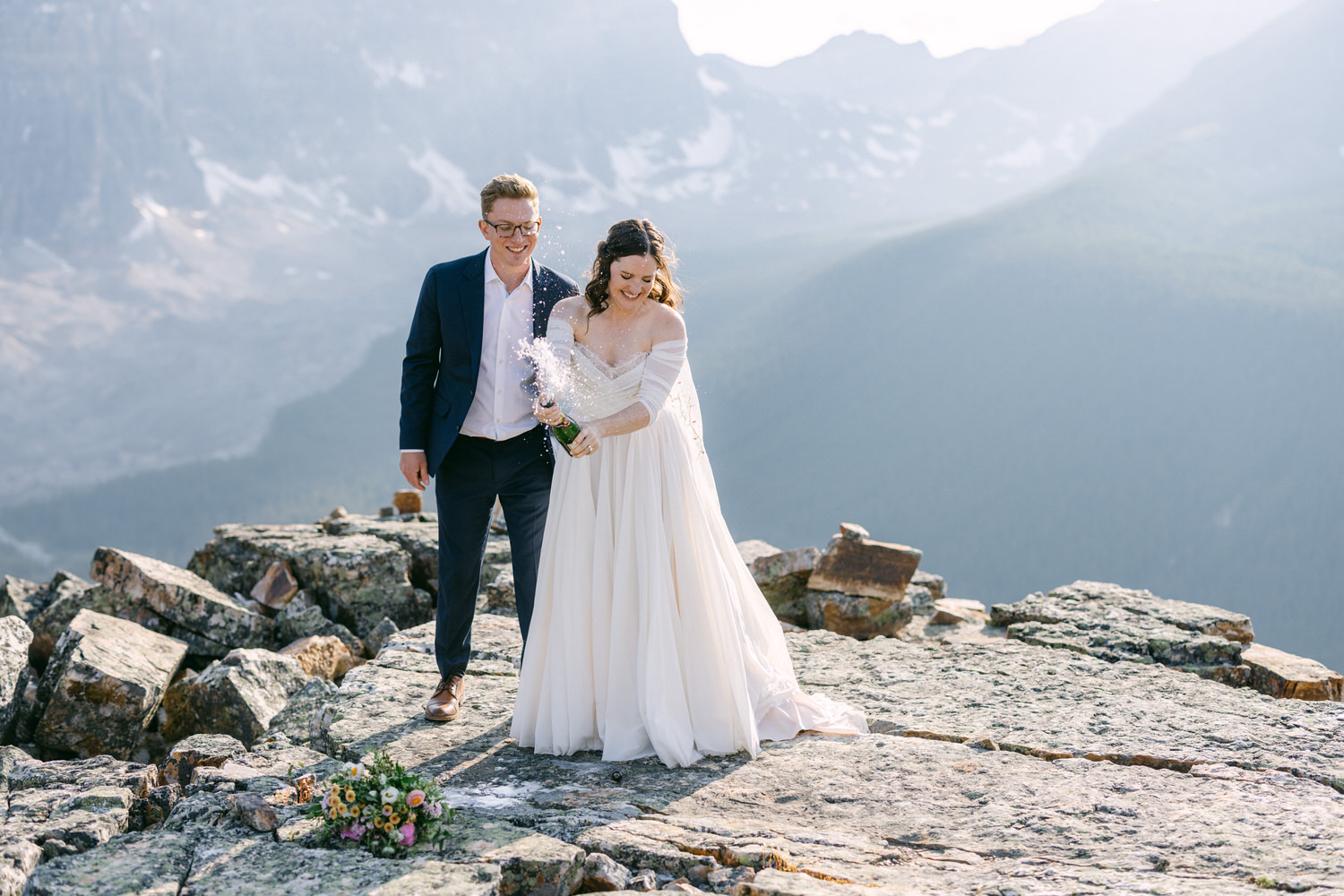 A joyful couple celebrates their wedding atop a rocky mountain, with one partner popping a champagne bottle as the other smiles, surrounded by stunning alpine scenery.