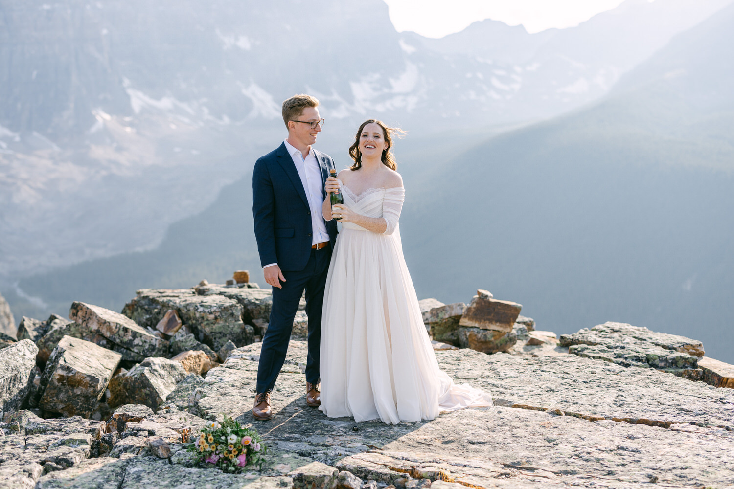 A happy couple stands on a rocky outcrop, with majestic mountains in the background, as the bride holds a champagne bottle and smiles.