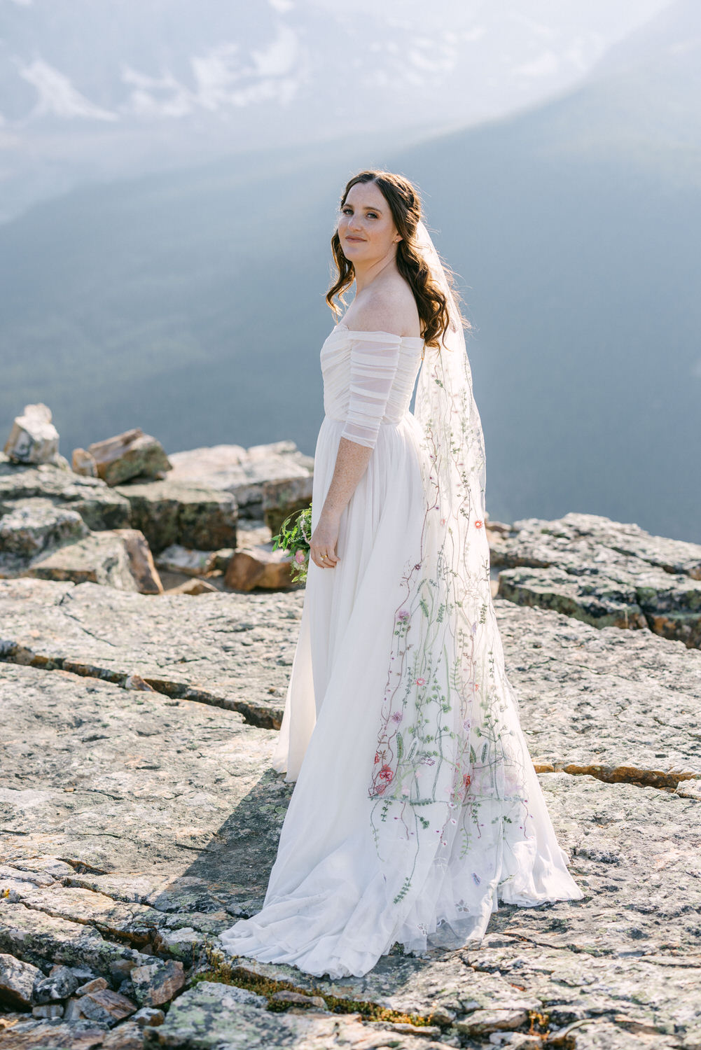 A bride in a flowing white gown with floral embroidery, standing on rocky terrain with a mountainous backdrop.