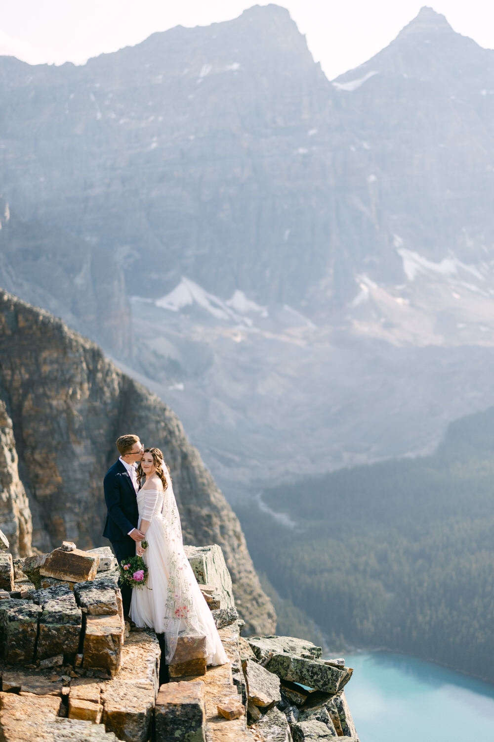 A couple sharing a romantic kiss on a rocky ledge with stunning mountain scenery and a serene lake in the background.