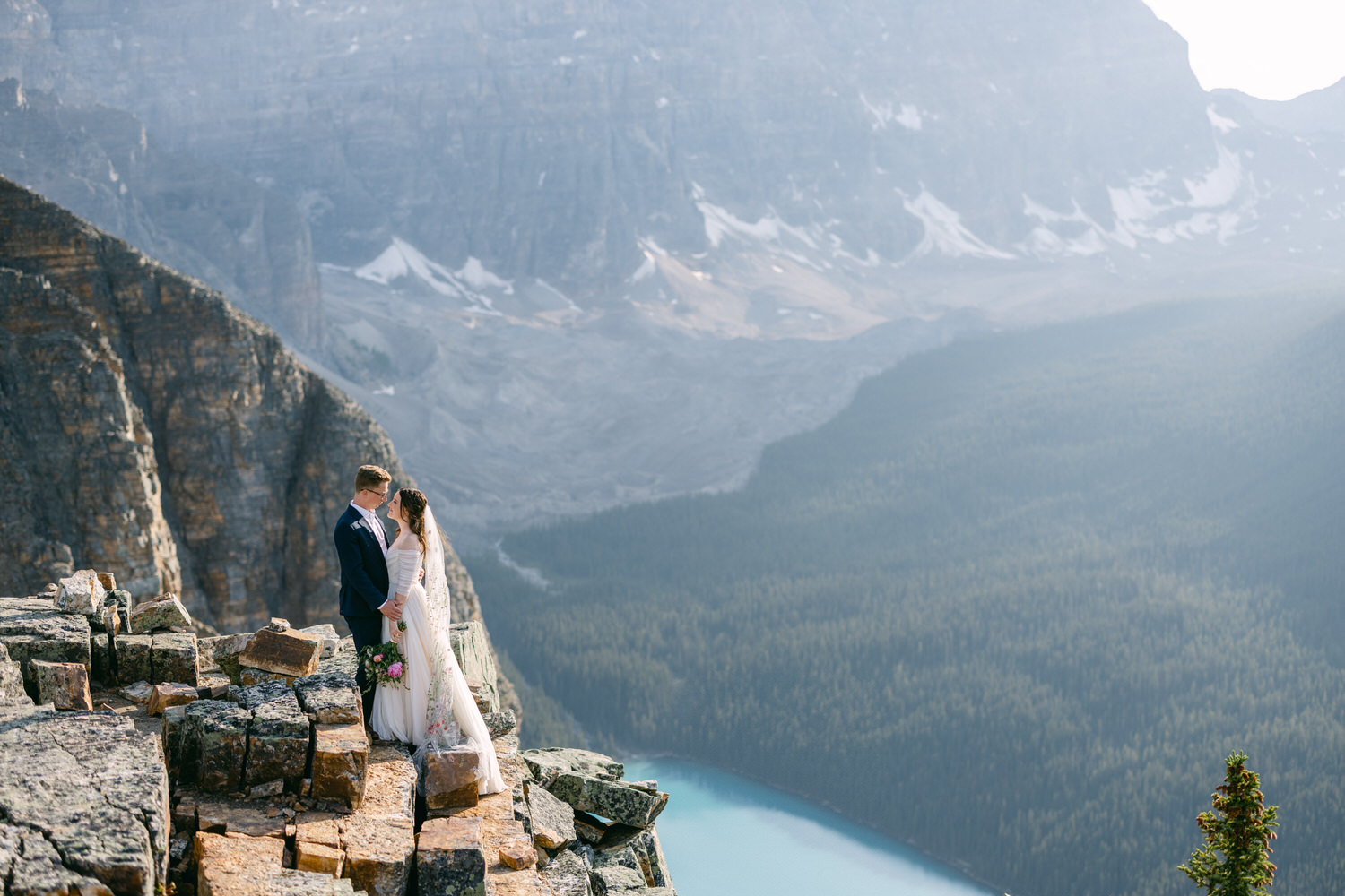 A couple sharing a romantic moment on a rocky ledge, surrounded by majestic mountain scenery and a serene lake in the background.