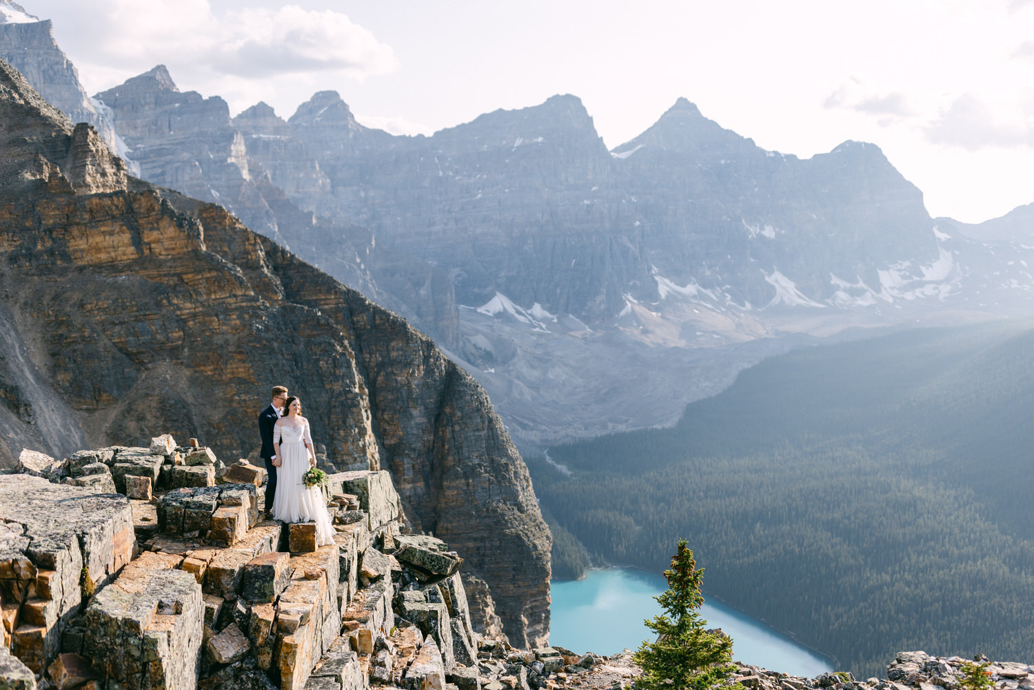 A couple in wedding attire stands on rocky terrain with a breathtaking mountain backdrop and a serene lake below.