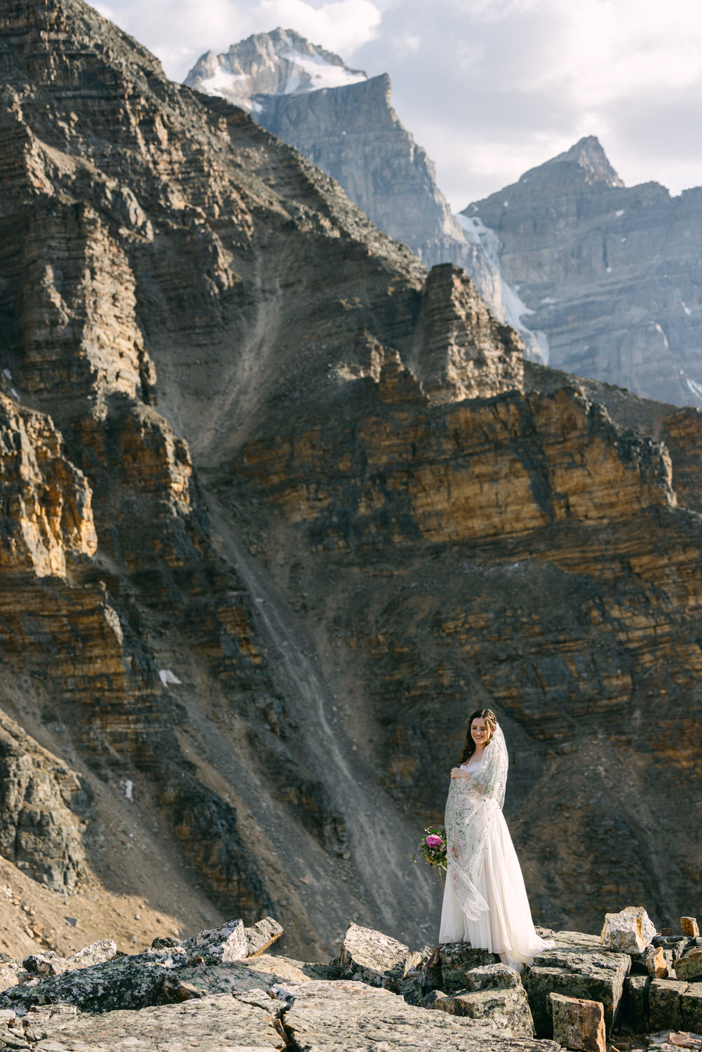 A bride in a flowing white dress and veil stands on rocky terrain against a stunning mountain backdrop, holding a bouquet of vibrant flowers.