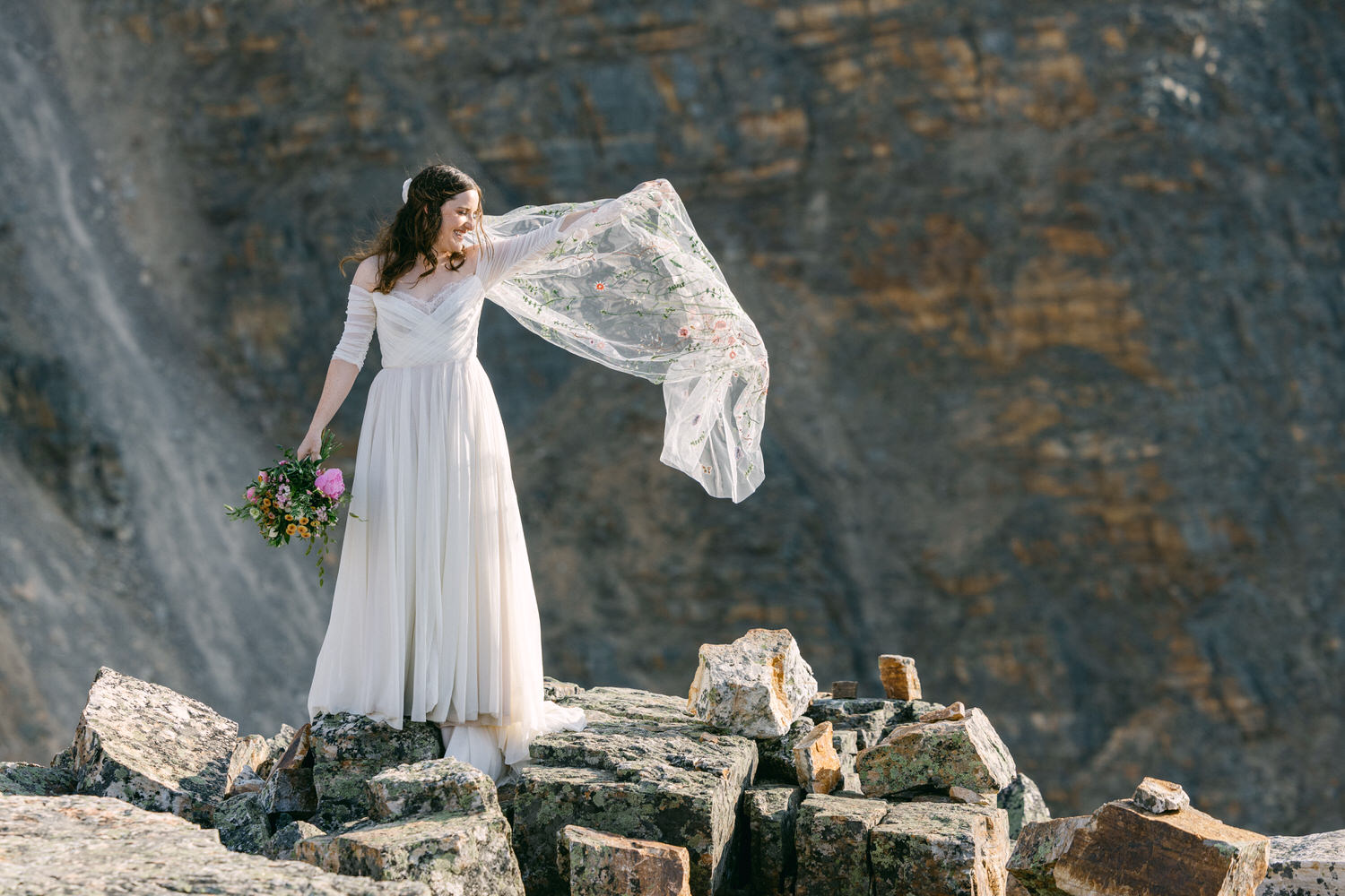 A joyful bride stands atop rugged rocks, gracefully holding a floral bouquet and a flowing, embroidered veil, with a stunning mountainous background.