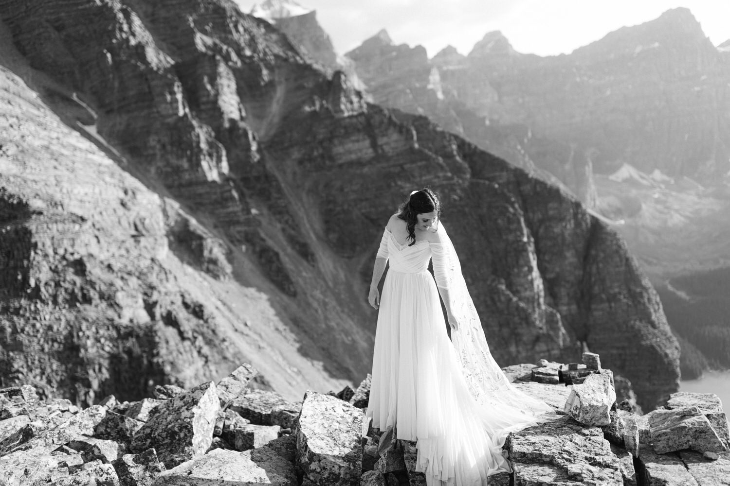 A bride in a flowing white gown and veil stands gracefully on rocky terrain against a backdrop of majestic mountains.