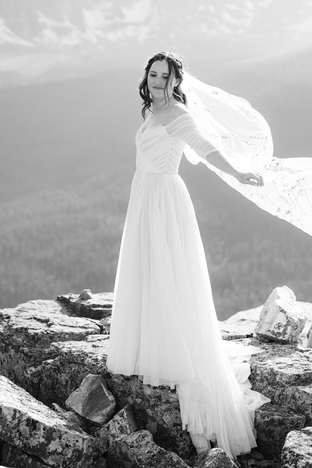 A bride with flowing hair stands gracefully on rocky terrain, her white dress and veil billowing in the wind against a misty mountain backdrop.