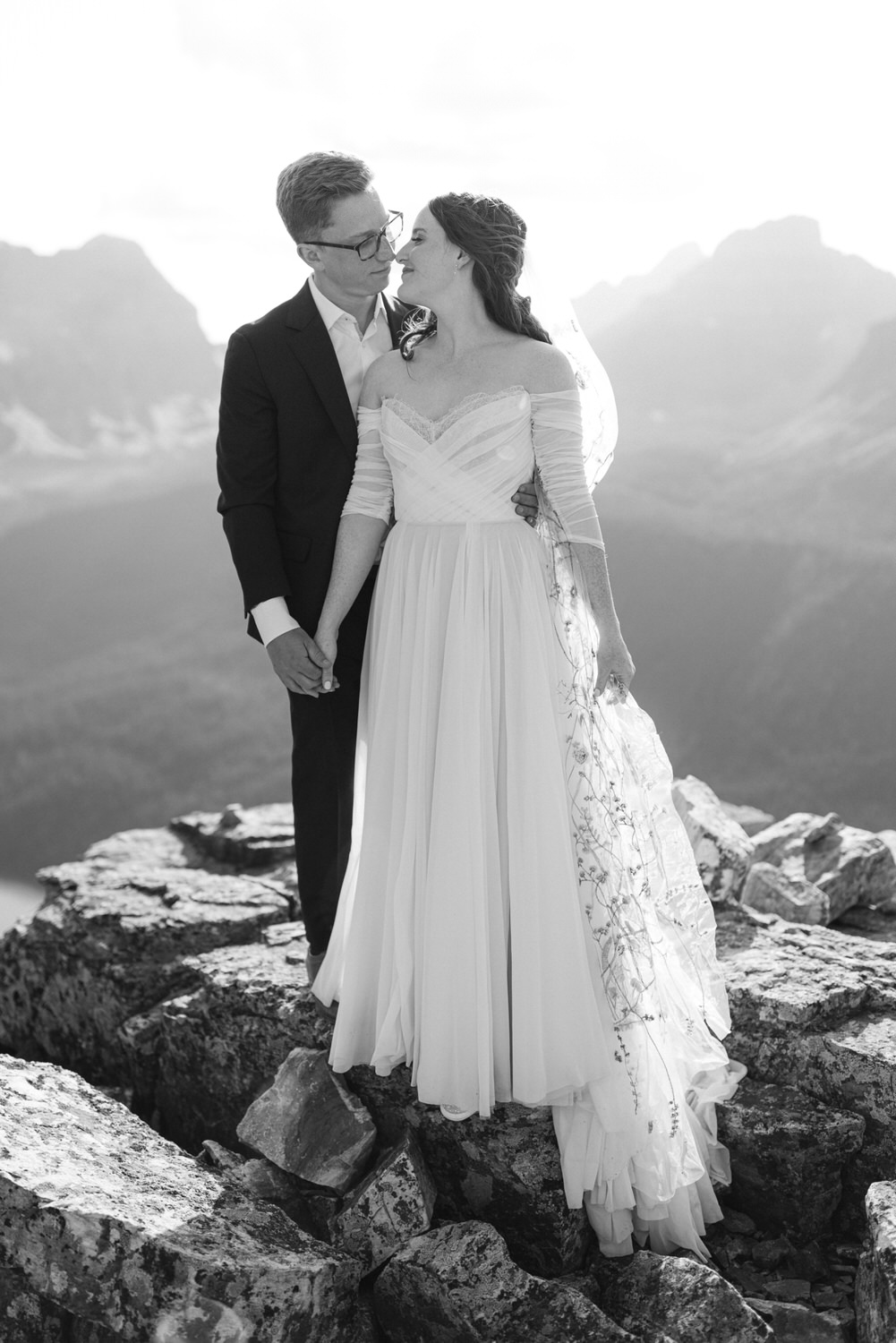 A couple sharing a tender gaze on a rocky mountain peak, dressed in wedding attire with a breathtaking backdrop of mountains and sky.