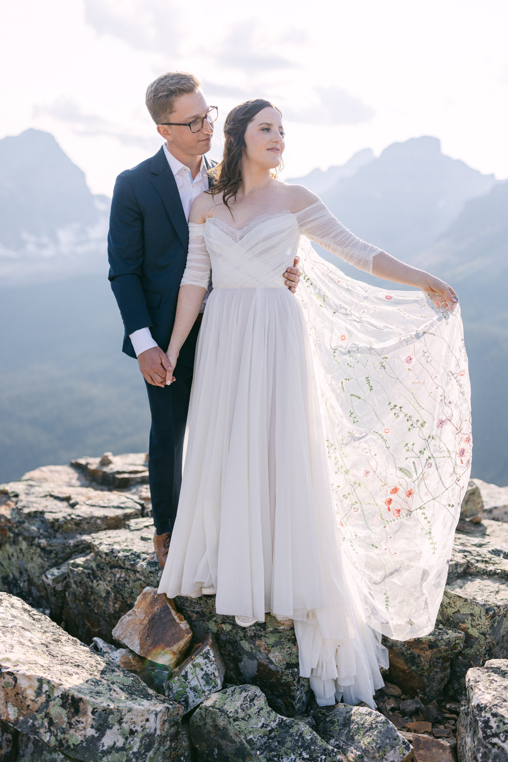 A couple stands in love on rocky terrain, surrounded by stunning mountains, as the bride wears a beautiful gown and delicate veil, embodying romance and adventure.