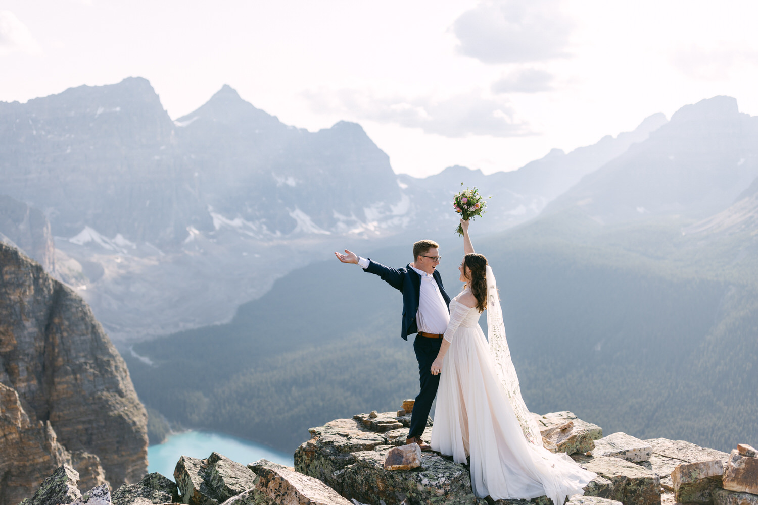 A couple joyfully celebrating their wedding on a rocky mountain peak, with stunning mountains and a turquoise lake in the background.