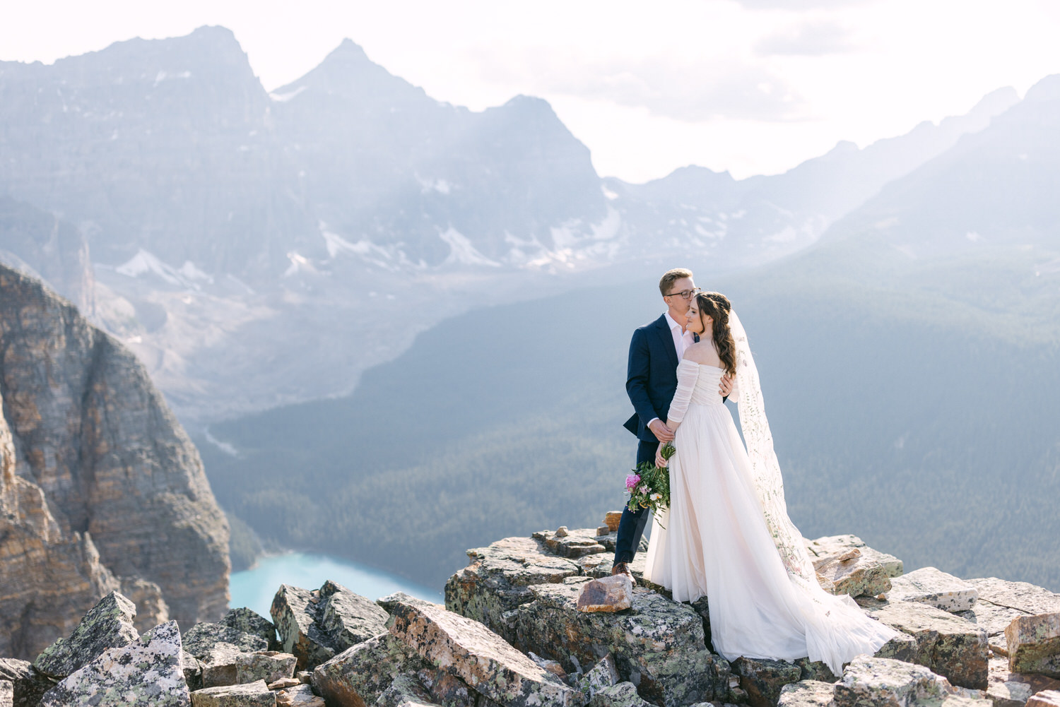 A couple sharing a romantic moment on a rocky mountaintop, surrounded by stunning landscapes of mountains and forests.