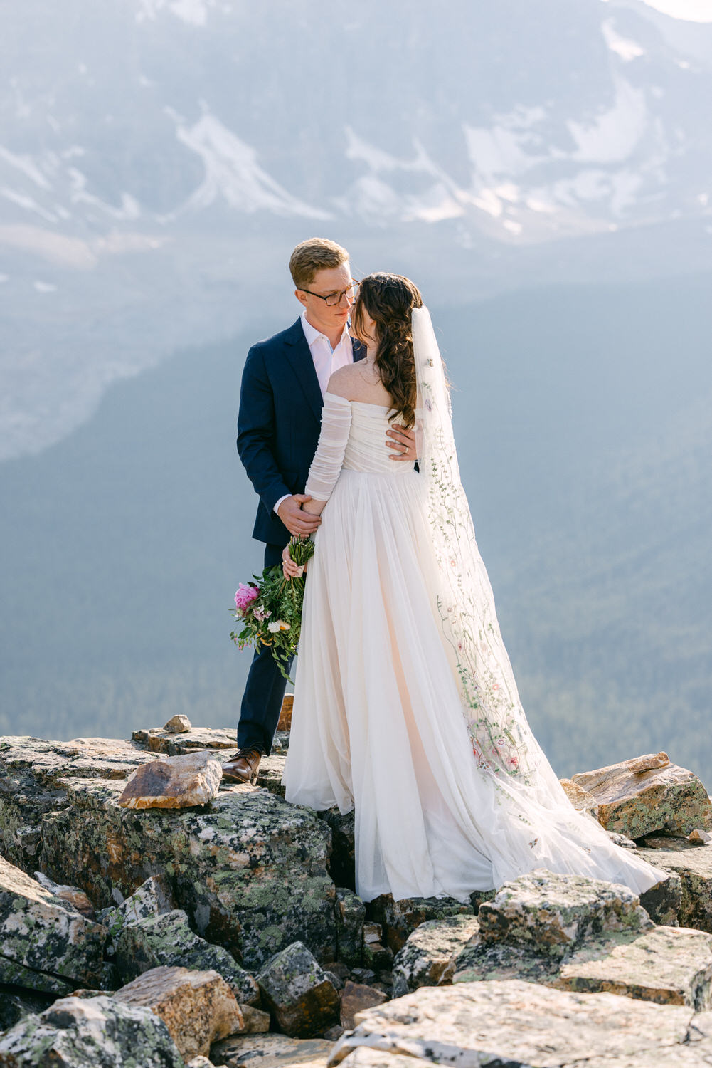 A couple shares a romantic moment on a rocky outcrop, framed by stunning mountain scenery and soft natural light.