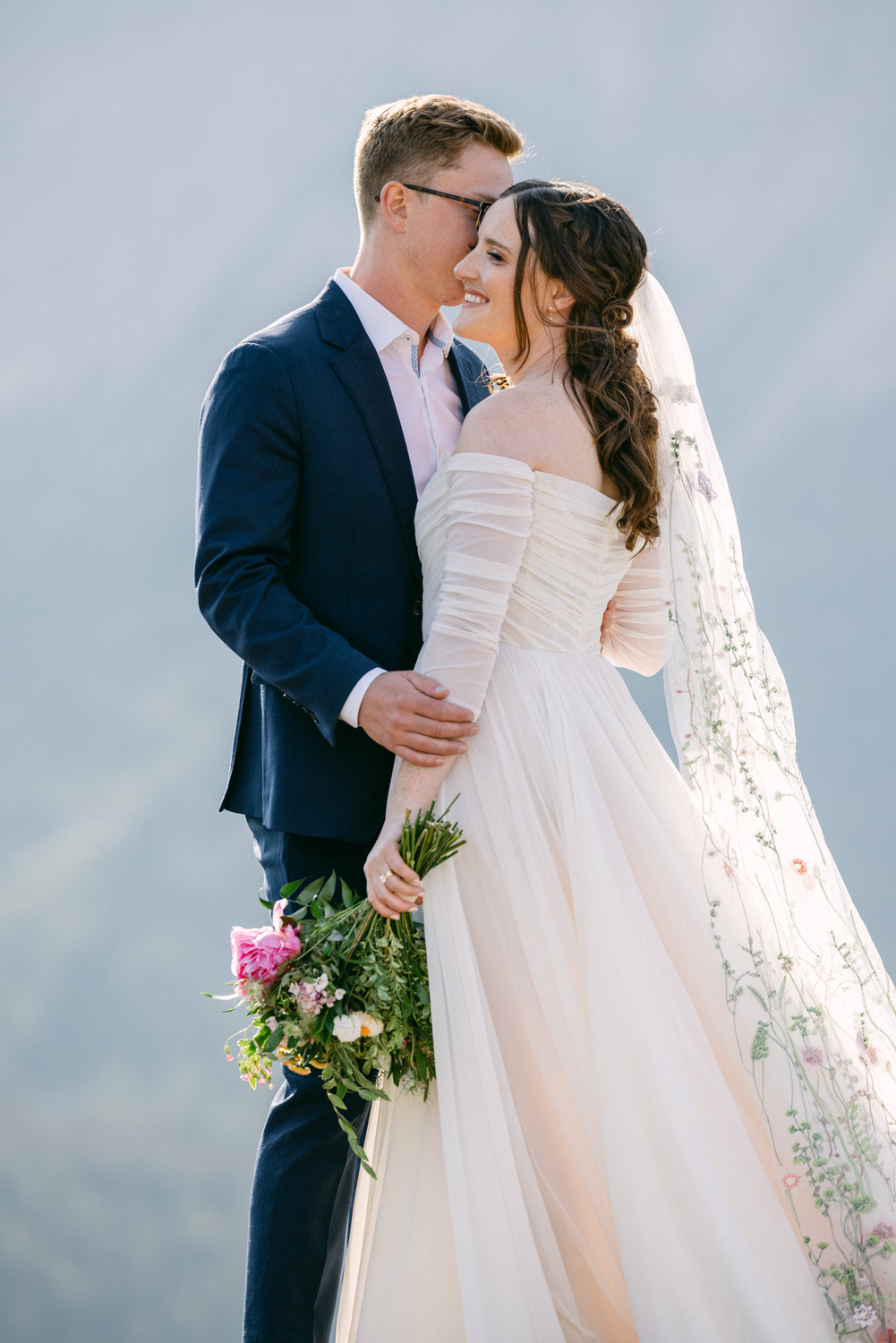 A bride and groom share a loving moment outdoors, with the bride holding a bouquet of flowers, dressed in an elegant off-shoulder wedding gown and a flowing veil, while the groom gently kisses her forehead.